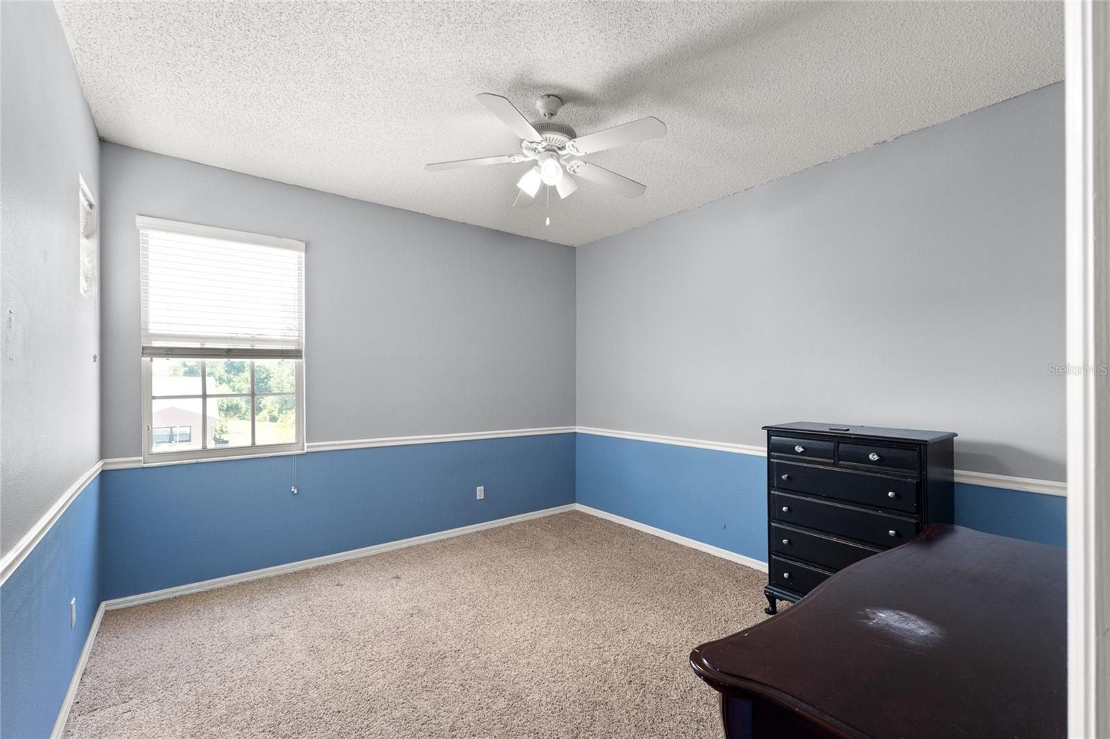 3rd second-floor bedroom with carpeted flooring, walk-in closet, neutral paint tones with chair rail detail, ceiling fan with light kit, and oversized window providing natural light.
