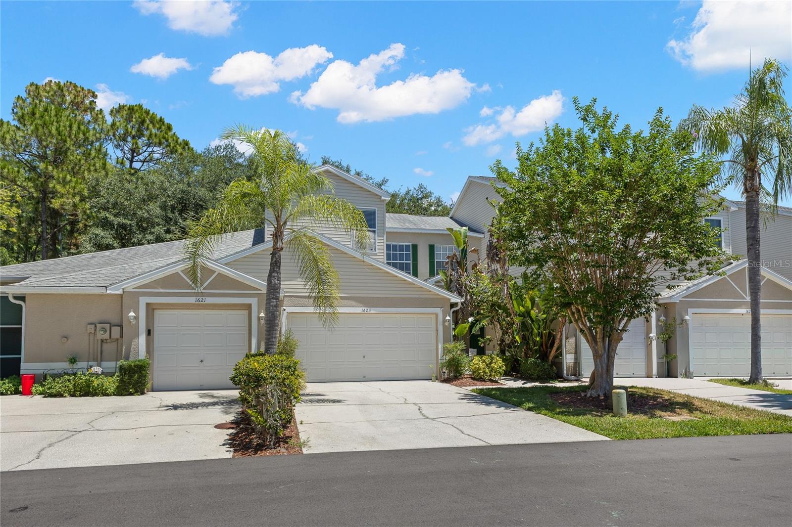 Exterior view of the home with a two-car garage, mature tropical landscaping, palm trees, and a welcoming front entry framed by lush greenery. Set within a well-maintained community with neighboring homes and tree-lined surroundings.