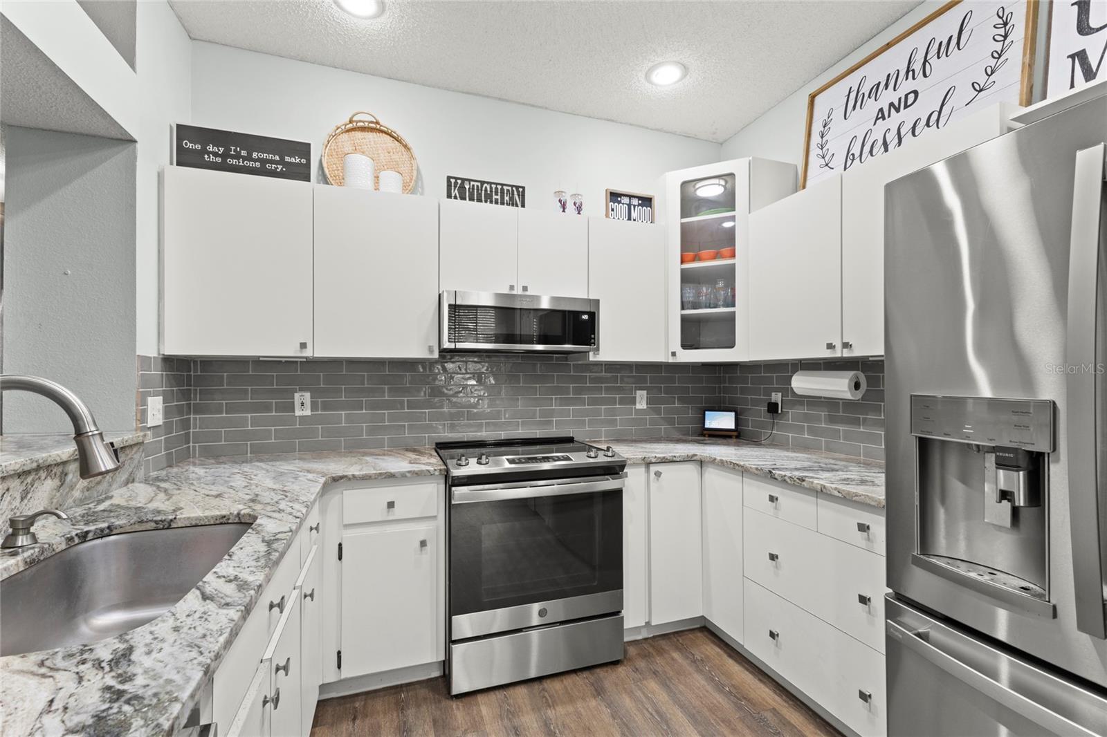 Kitchen with white shaker-style cabinetry, granite countertops, stainless steel appliances, gray subway tile backsplash, recessed lighting, and wood-look laminate flooring. Upper display cabinetry and extended-height wall space enhance the kitchen’s open, airy feel.