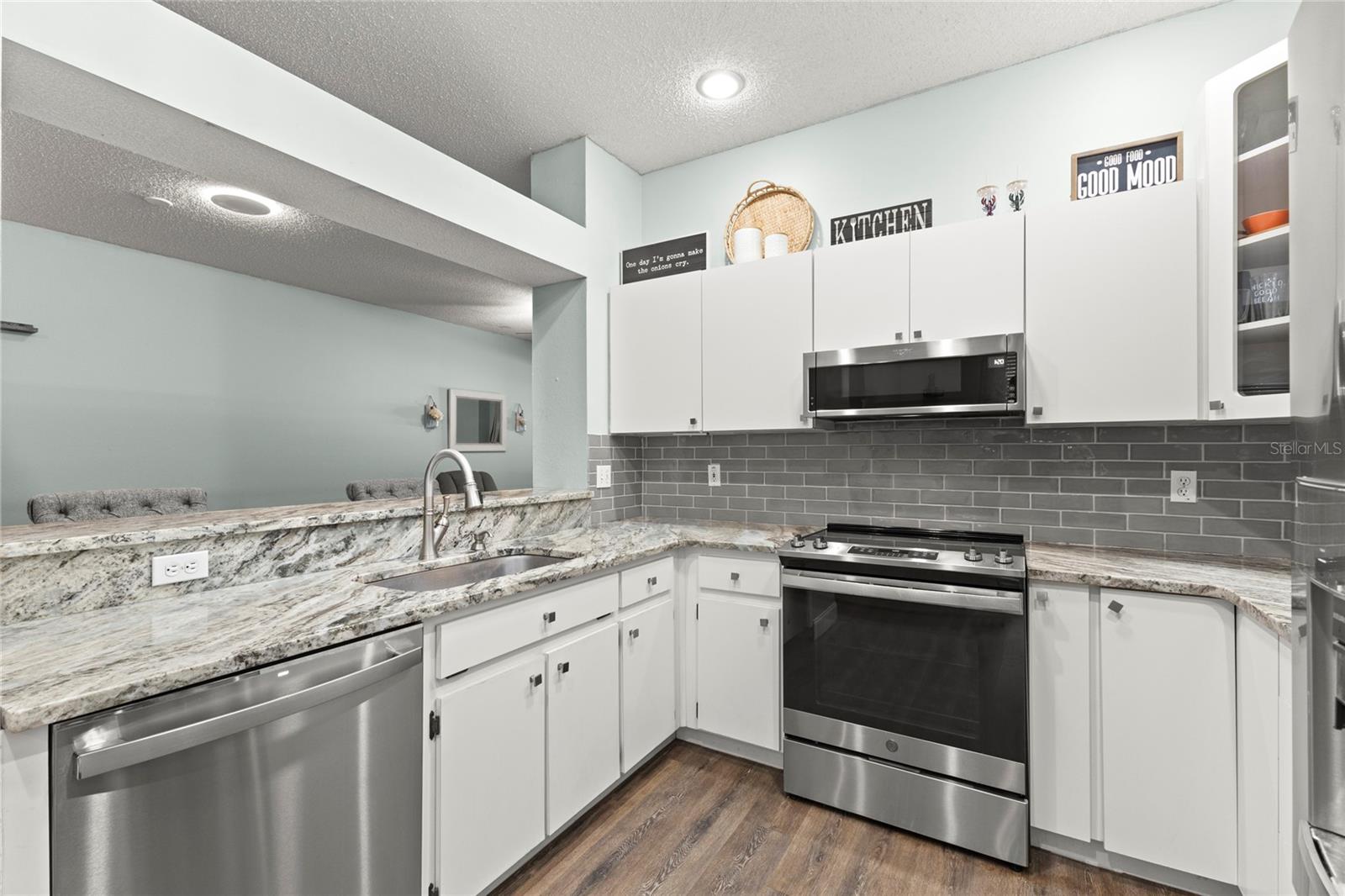 Kitchen with granite countertops, white shaker-style cabinetry, stainless steel appliances, a gray subway tile backsplash, recessed lighting, and a breakfast bar with pendant-ready overhang seating. The raised passthrough opening creates connection to the adjoining living and dining areas while maintaining defined spaces.