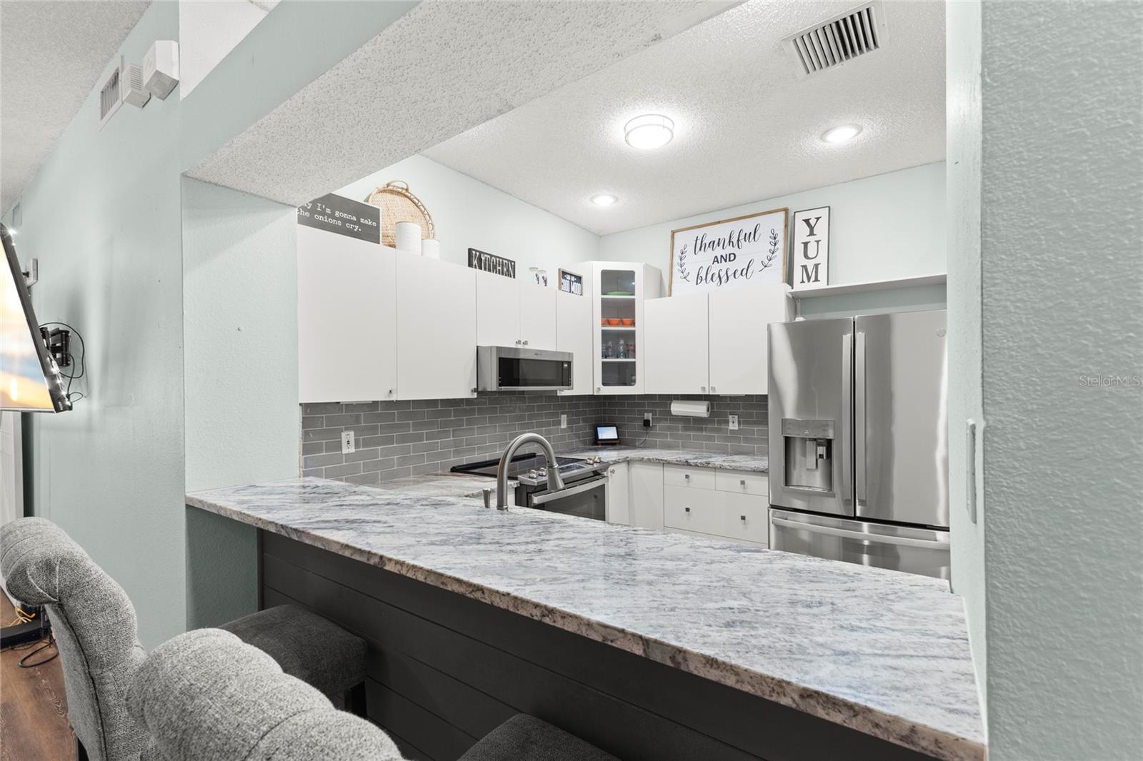 Oversized breakfast bar with granite countertops and bar seating overlooking the kitchen, featuring white shaker-style cabinetry, stainless steel appliances, recessed lighting, and a gray subway tile backsplash. The raised passthrough design creates separation while maintaining connection to the adjoining living spaces.