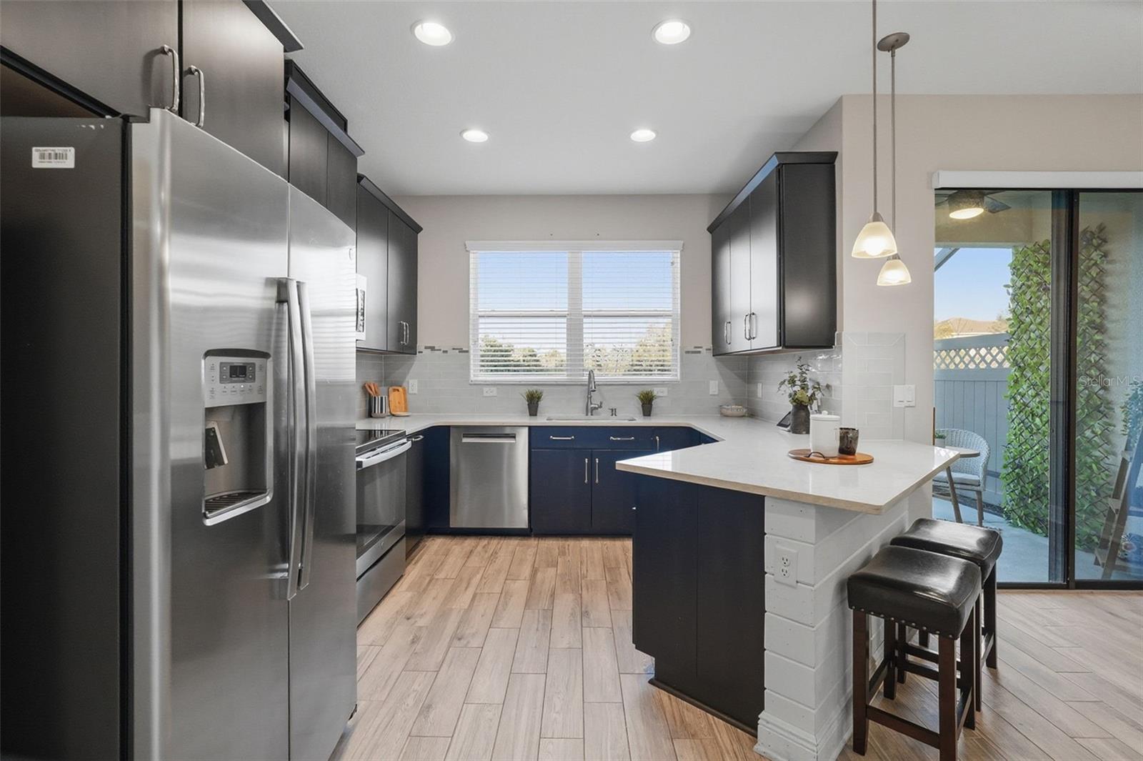 Updated kitchen featuring sleek white quartz countertops, stainless steel appliances, dark shaker-style cabinetry, recessed lighting, and a spacious breakfast bar perfect for casual dining and entertaining. Large windows bring in natural light, while the modern backsplash and wood-look tile flooring add warmth and style throughout