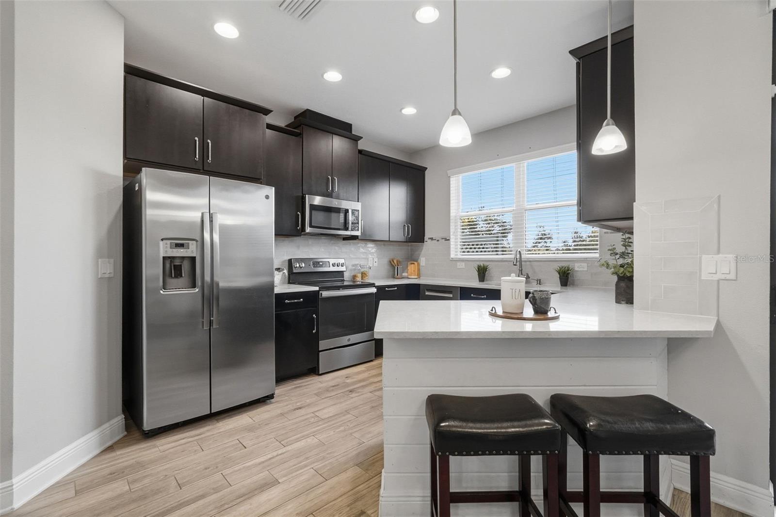 Updated kitchen featuring sleek white quartz countertops, stainless steel appliances, dark shaker-style cabinetry, recessed lighting, and a spacious breakfast bar perfect for casual dining and entertaining. Large windows bring in natural light, while the modern backsplash and wood-look tile flooring add warmth and style throughout