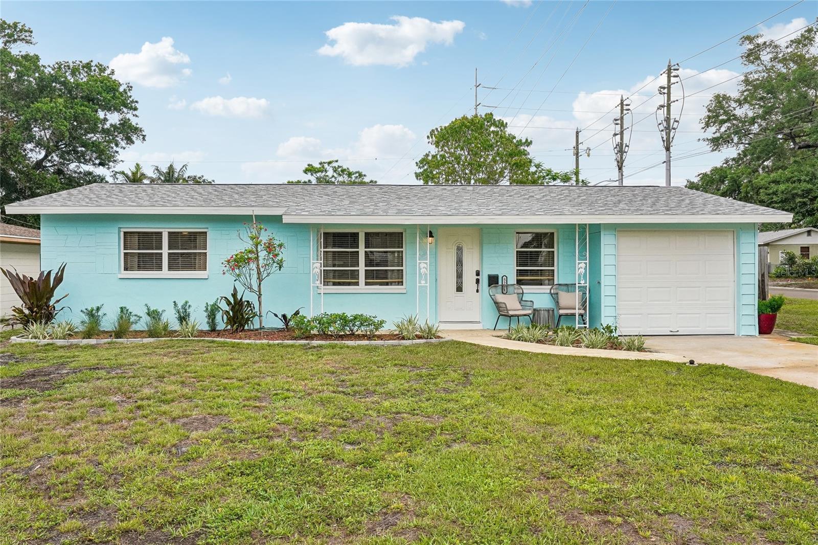 Front Exterior — Concrete block ranch-style home with a fresh aqua exterior, white garage door, and covered front entry with seating area. Mature landscaping, tropical plantings, and a lush front lawn add to the inviting curb appeal.