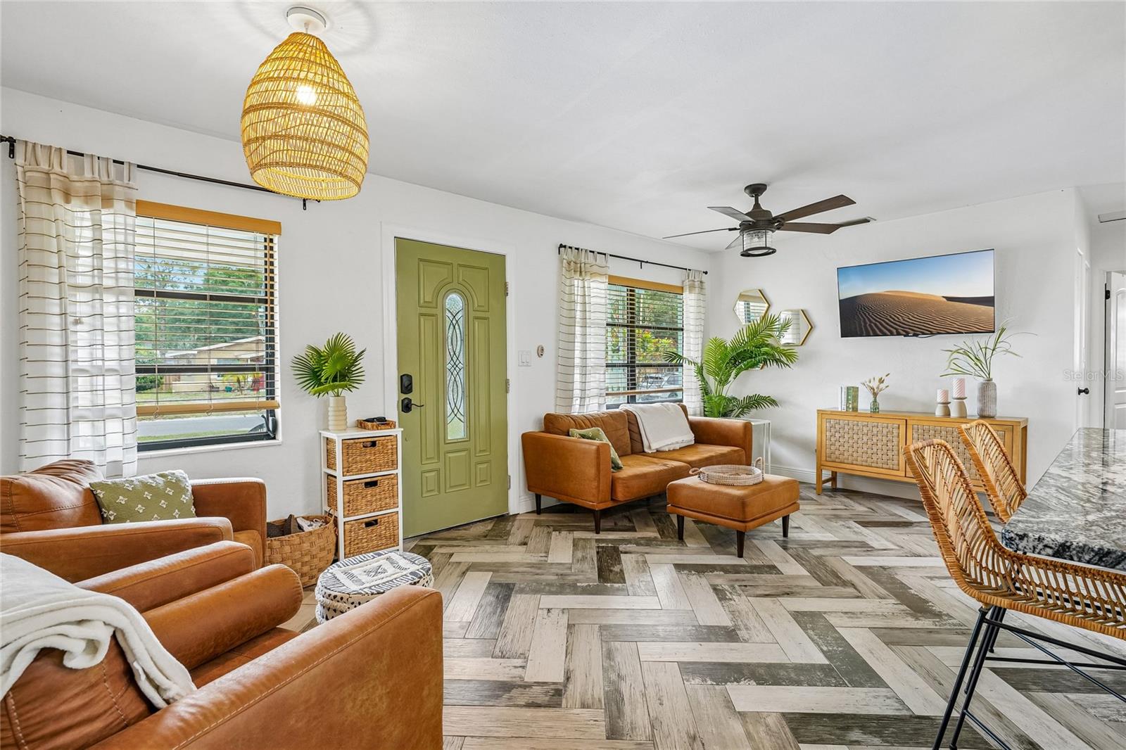 Living Room — Open living area with herringbone-pattern tile flooring, a woven rattan pendant light, and a glass-paneled entry door. Multiple windows bring in natural light, and the layout flows open toward the kitchen and dining areas.