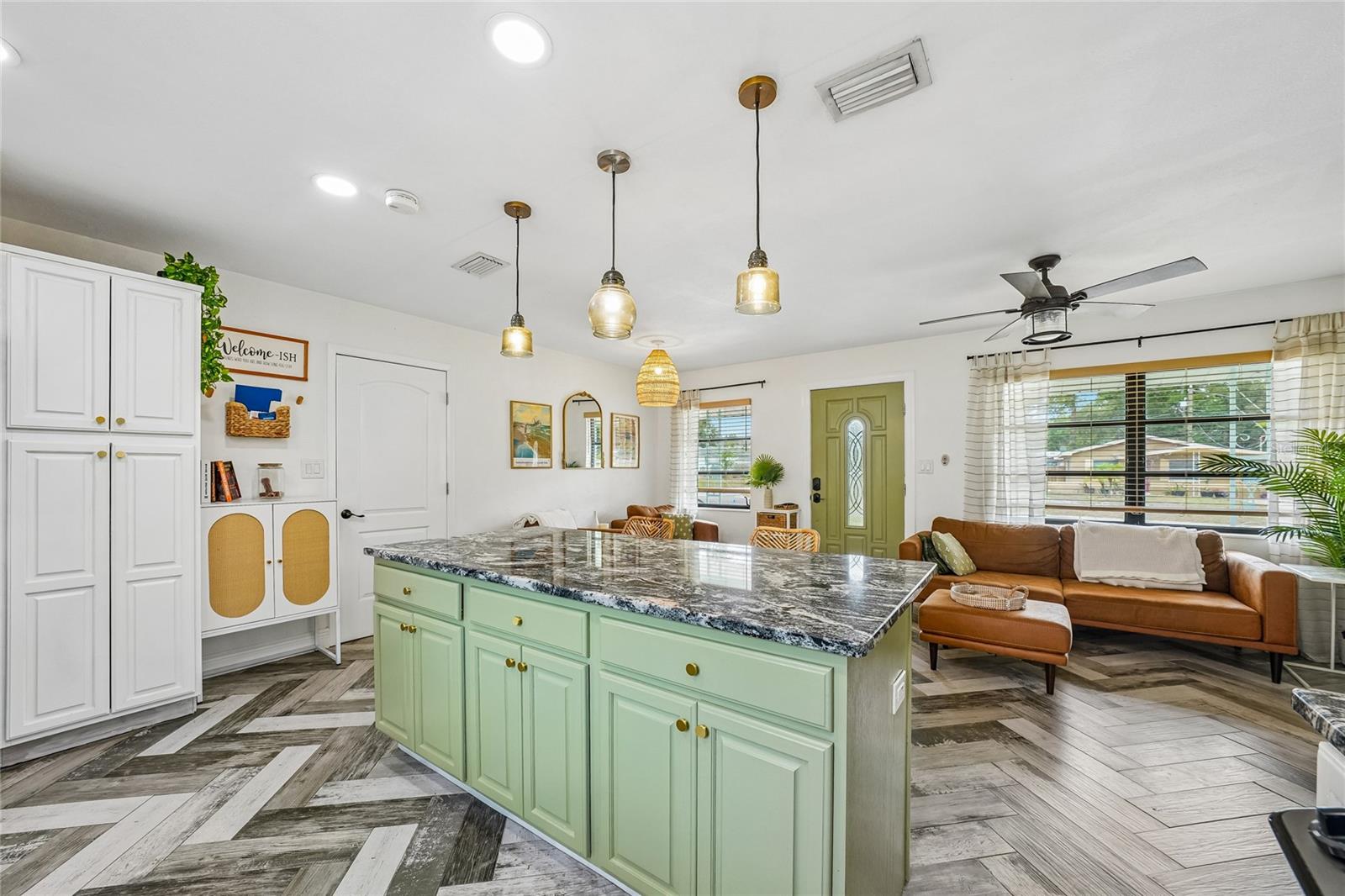 Kitchen — Green beadboard island with dark granite countertop, brass hardware, and amber glass pendant lights. Herringbone tile flooring runs through the kitchen and into the open living area.
