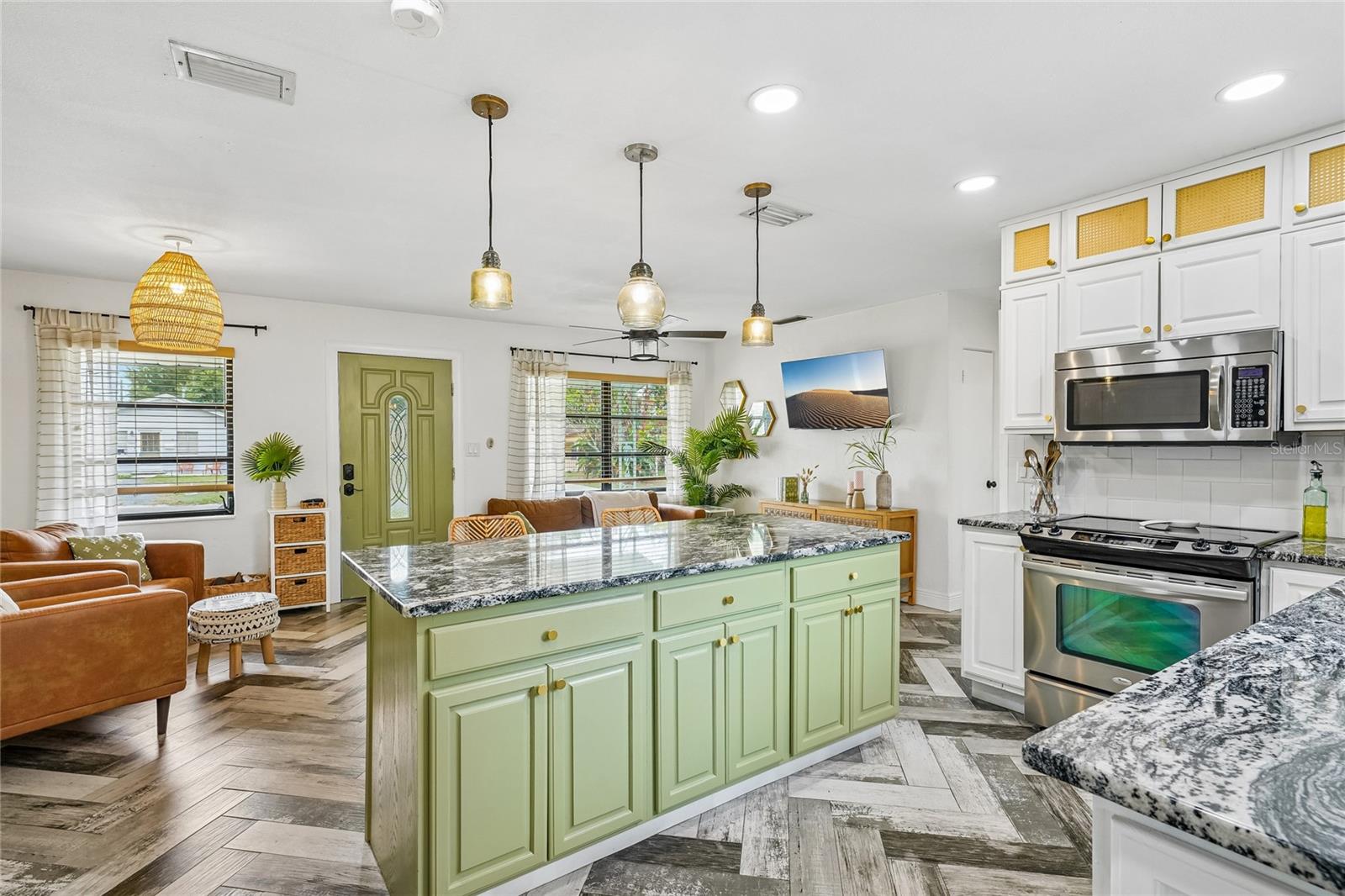 Kitchen — Full view of the green center island with dark granite countertop, brass hardware, and three amber glass pendant lights. White upper cabinetry with rattan inserts, stainless steel range and built-in microwave, and subway tile backsplash visible.