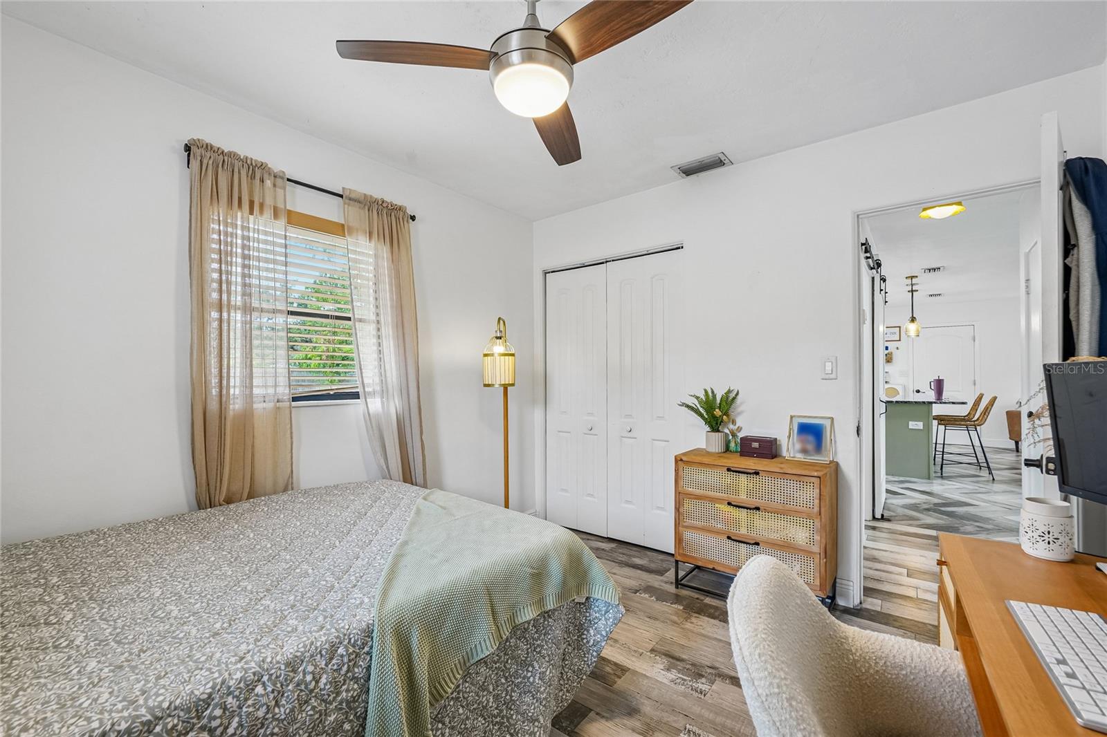 Bedroom 1 — View showing the reach-in closet with bi-fold doors, tile flooring, ceiling fan, and open sightline to the kitchen. Window with blinds and sheer curtains.
