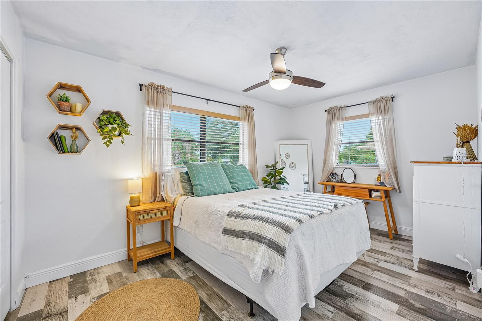Bedroom 2 — Bedroom with tile flooring, ceiling fan, and two windows with blinds and sheer curtains. Hexagonal wall shelves and natural light throughout.