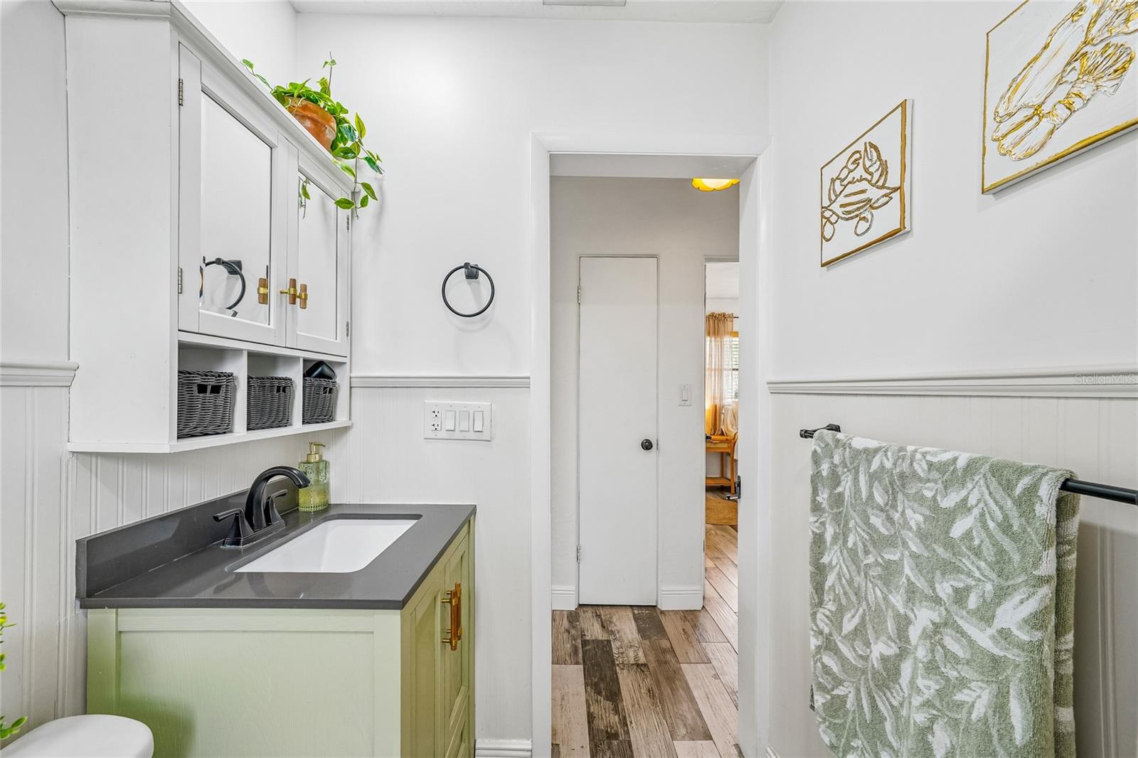 Bathroom — Bathroom with green vanity cabinet, dark countertop, undermount sink, matte black faucet, and white upper wall cabinet with open storage cubbies. Beadboard wainscoting and tile flooring throughout.