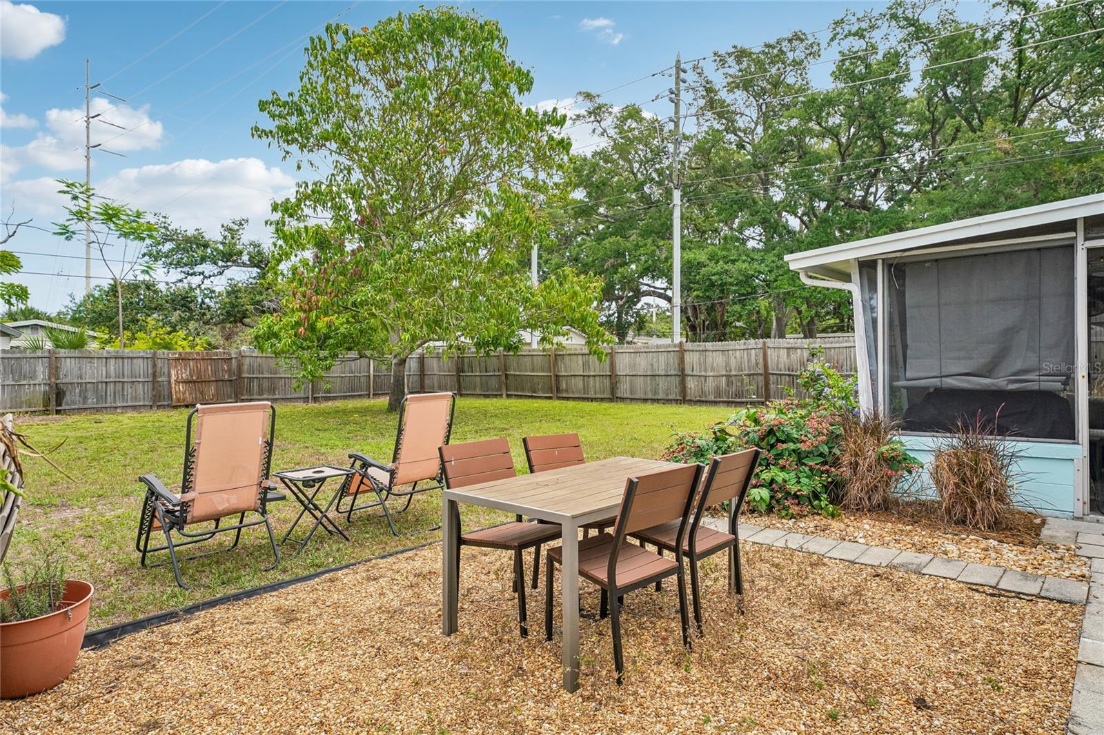 Backyard — Fenced backyard with gravel patio, mature trees, and open lawn. Screened lanai visible at rear of home.