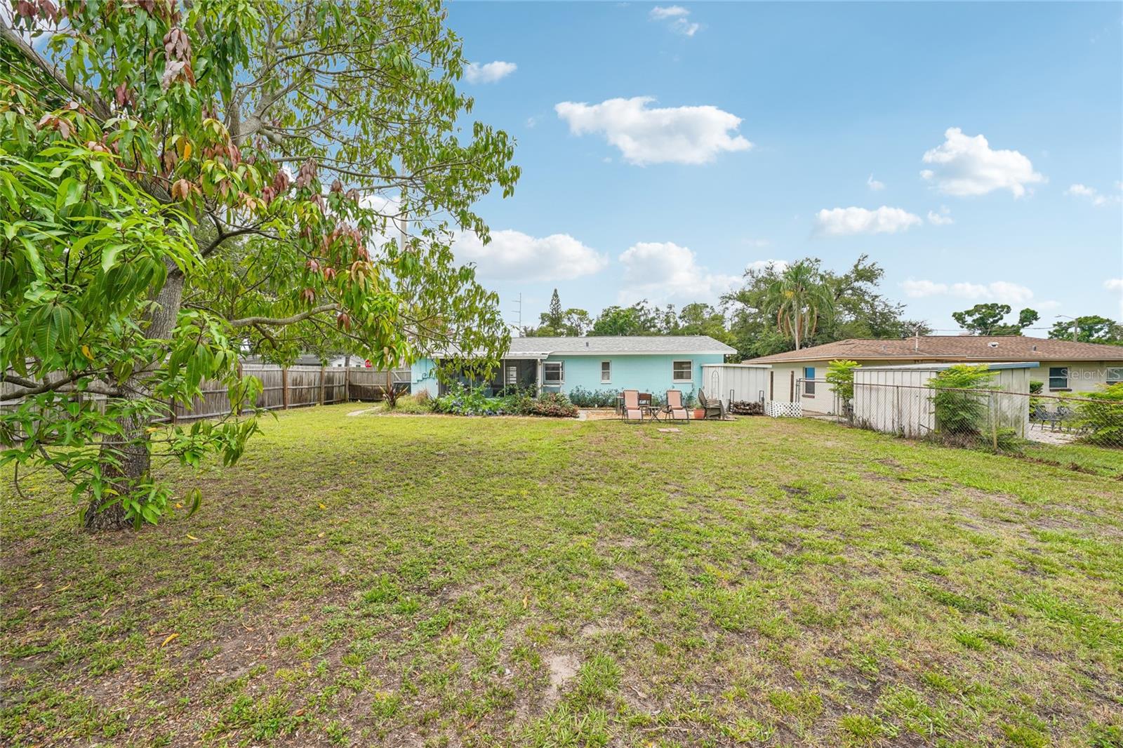 Backyard — Wide view of the fully fenced backyard with open lawn, mature shade trees, and the rear exterior of the home visible. Gravel patio seating area at rear of home.
