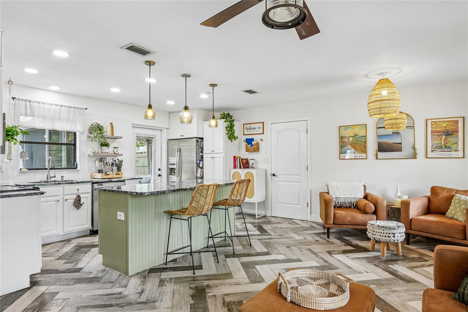 Kitchen / Living Room Wide-angle view showing the open-concept layout connecting the kitchen island to the living area, with herringbone tile flooring running throughout. Green beadboard island with dark granite countertop, three amber glass pendant lights, and recessed lighting above.