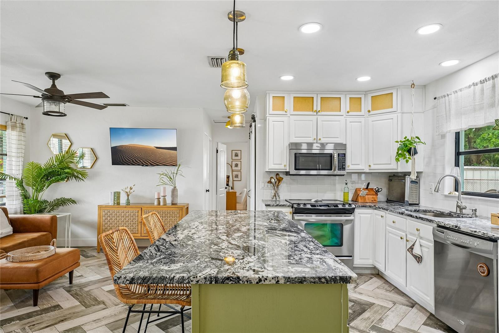 Kitchen — View from the island looking toward the cooking area, with white cabinetry, subway tile backsplash, stainless steel range and dishwasher, and dark granite countertops. Open sightline to the living room beyond, with herringbone tile flooring throughout.