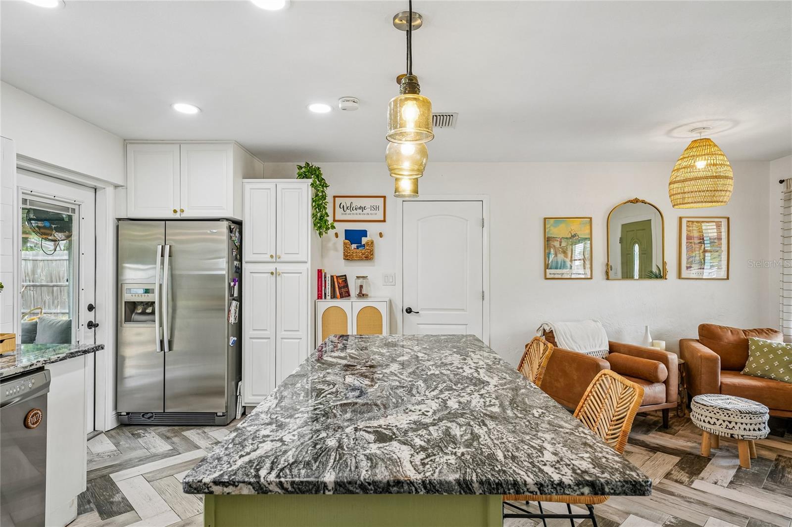 Kitchen — View from the island toward the refrigerator wall, with stainless steel French door refrigerator, tall white pantry cabinetry, and dark granite island countertop. Open flow to the living area.