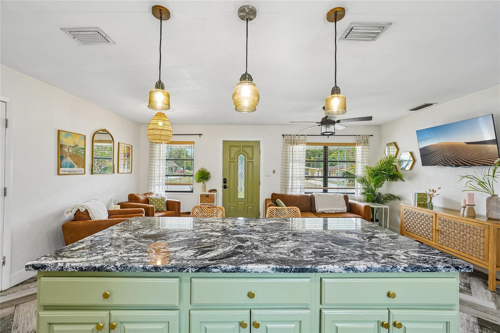 Kitchen / Living Room — View from behind the island looking toward the living room and front entry, with the full granite island, green beadboard cabinetry and three amber glass pendant lights. Herringbone tile flooring runs continuously through both spaces.
