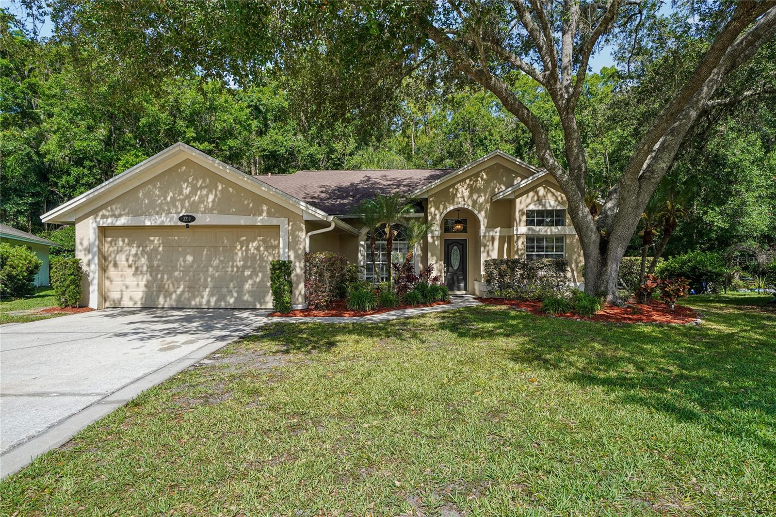 Exterior front — Stucco exterior with a two-car garage, arched entry with decorative pendant light, and mature oak tree canopy framing the front yard. Manicured landscaping with tropical plantings and red mulch beds line the driveway and entry walk.