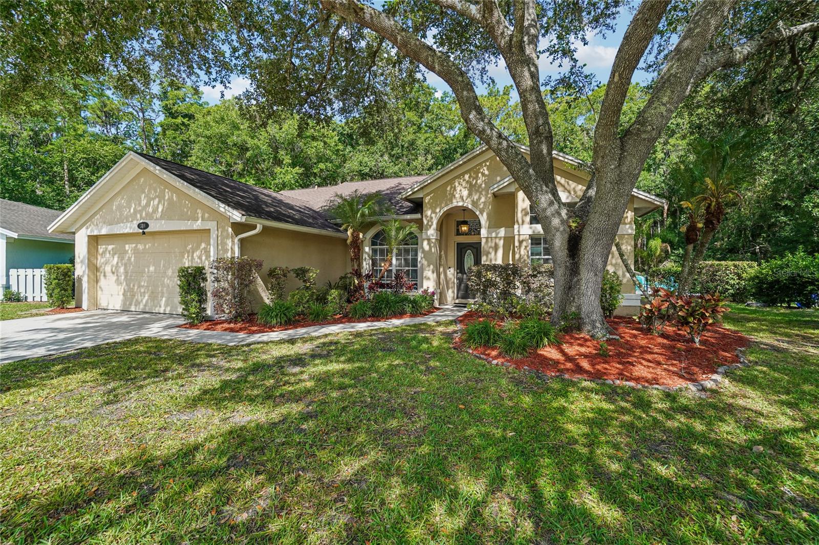 Exterior front — Full front elevation of the stucco home with a two-car garage, arched entry, and mature tree canopy providing natural shade across the front lawn. Tropical plantings and red mulch accent the landscaped beds.
