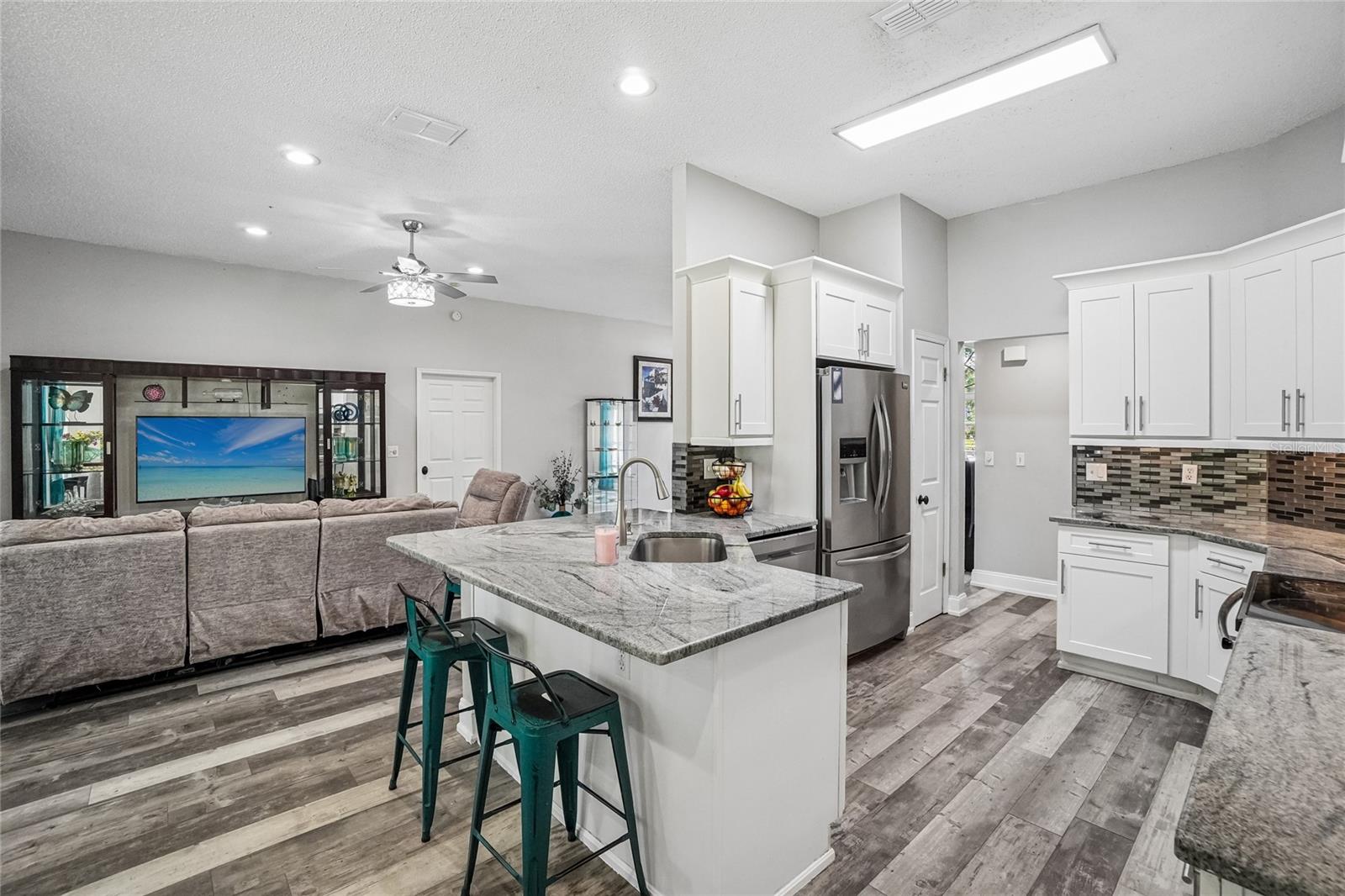 Kitchen / great room — Kitchen peninsula with granite countertop and single-basin sink opens directly to the great room. White shaker cabinetry, stainless steel refrigerator, and mosaic tile backsplash are visible, with the living area and ceiling fan beyond.