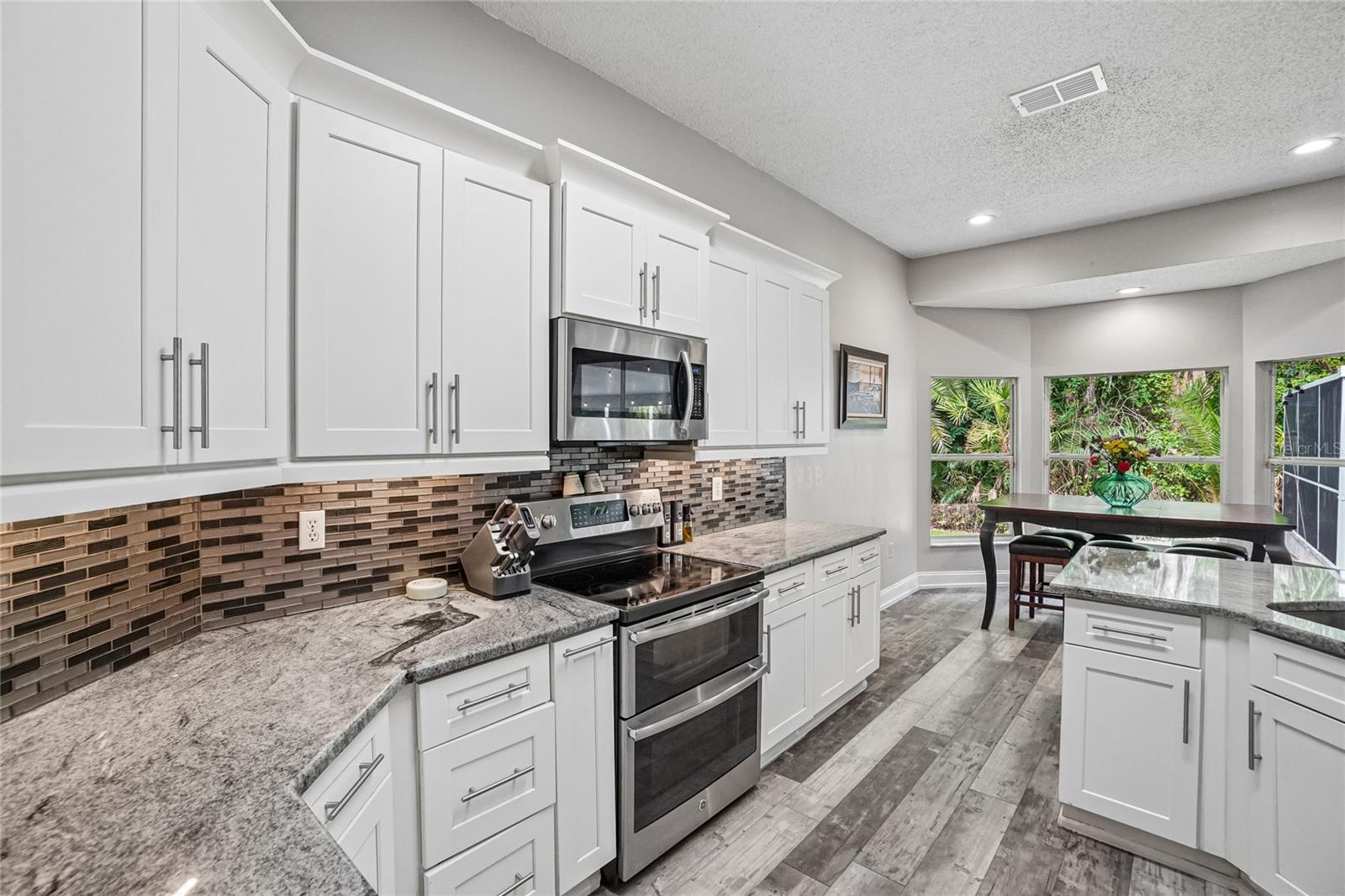 Kitchen — White shaker cabinetry with granite countertops, mosaic tile backsplash, and stainless steel double-oven range and over-the-range microwave. Sightlines extend to the dining area and backyard conservation through large windows.