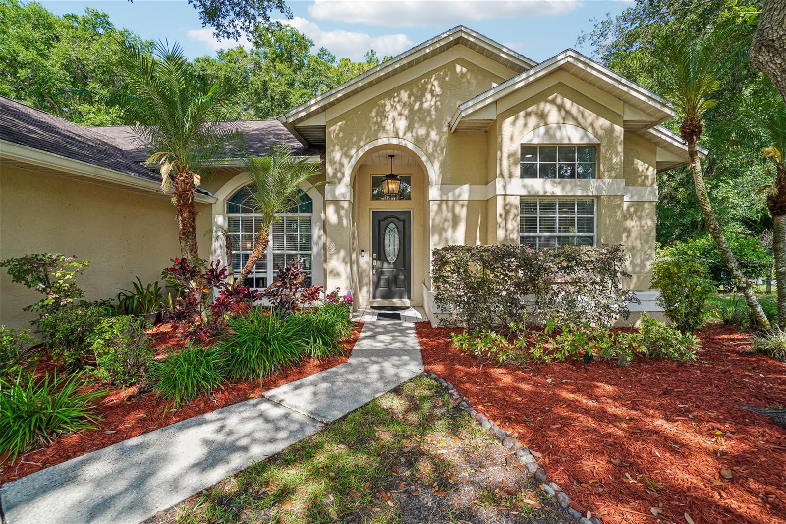 Front entry / exterior close-up — Arched covered entry with a decorative oval glass front door, lantern pendant light, and walkway bordered by tropical landscaping and red mulch.