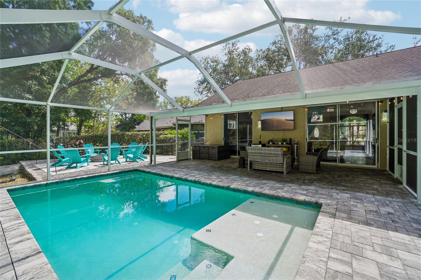 Pool / screened enclosure — In-ground pool with pavered pool deck and a covered lanai with outdoor seating area. The paver fire pit area with seating is visible beyond the pool cage, backed by mature oak canopy and a fenced yard.