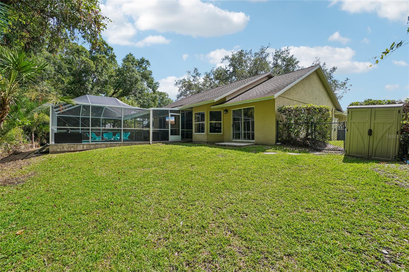 Backyard — Rear exterior view showing a spacious grass lawn, screened pool enclosure, and a utility shed with gated side yard access. Mature oak and palm canopy frame the perimeter of the property.