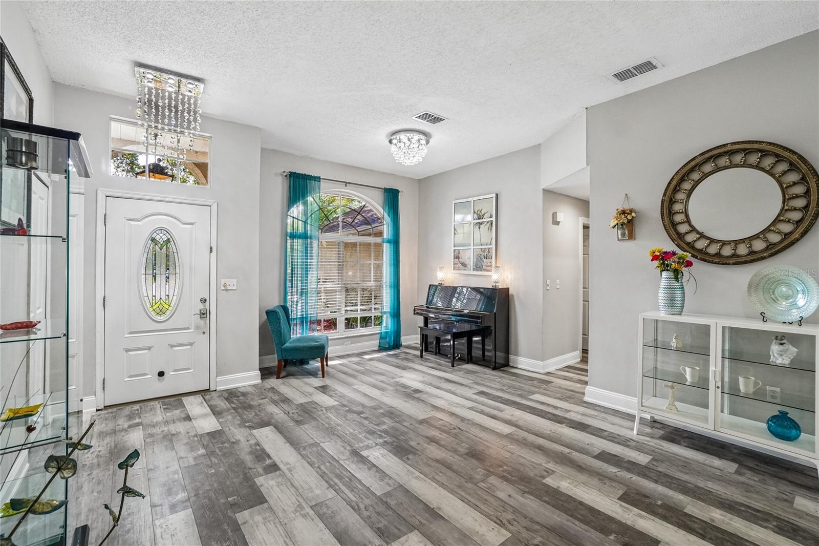 Foyer — Open foyer with luxury vinyl plank flooring, a glass oval insert in the front door, and two crystal flush-mount light fixtures. Flows directly into the flex room and main living areas.