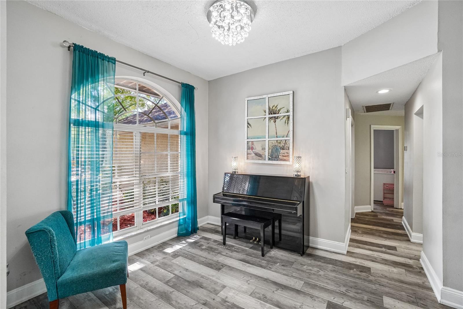 Flex room / sitting room — Light-filled flex room with luxury vinyl plank flooring, a crystal flush-mount light fixture, and a large arched window with plantation blinds. Opens to the hallway.