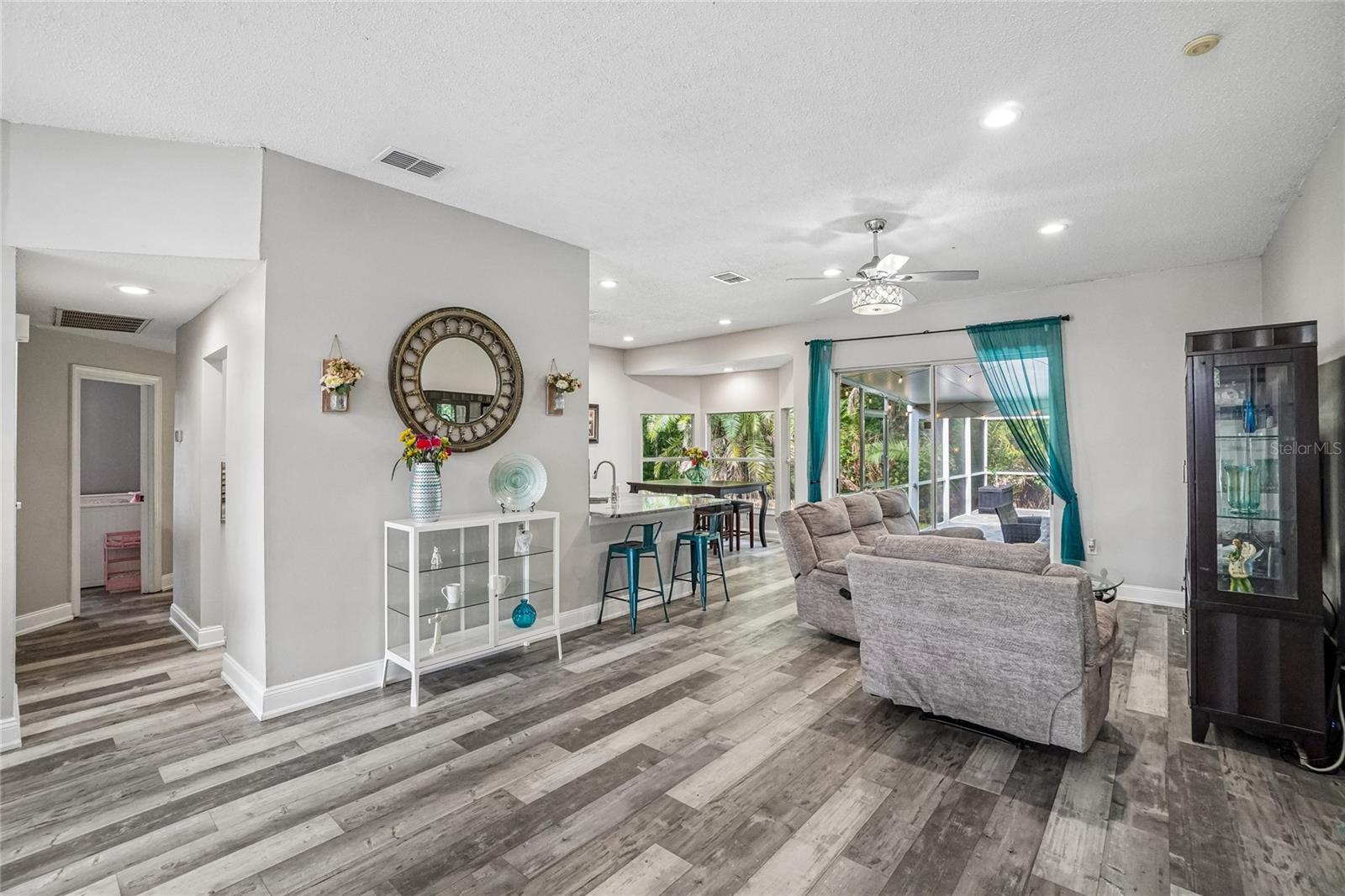 Living room / great room — Open great room with luxury vinyl plank flooring, recessed lighting, and a ceiling fan with decorative light kit. Sightlines carry through to the kitchen peninsula and sliding glass doors leading to the screened lanai.