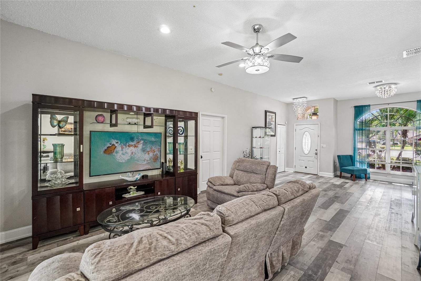 Living room — Great room with luxury vinyl plank flooring, ceiling fan with decorative light kit, and a view through to the foyer entry. Large arched window with blinds brings in natural light from the front of the home.