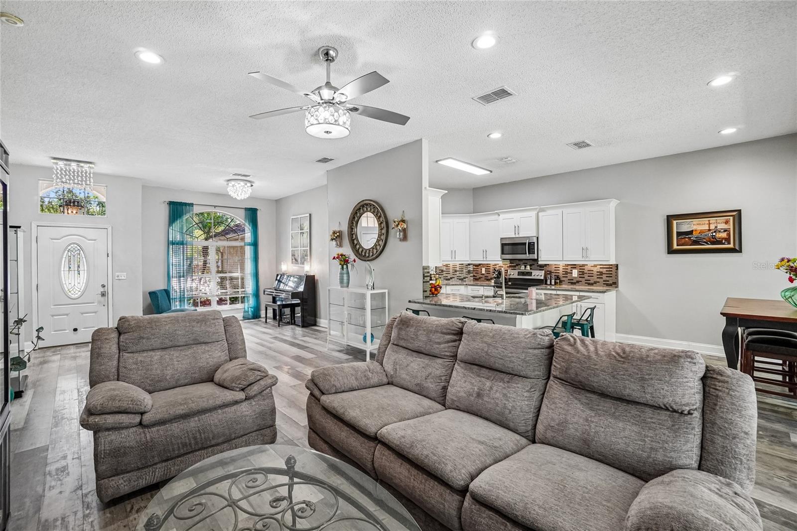 Living room — Great room with luxury vinyl plank flooring, ceiling fan with decorative light kit, and an open view to the foyer and arched front window. White shaker cabinetry and a tile backsplash are visible in the adjacent kitchen.