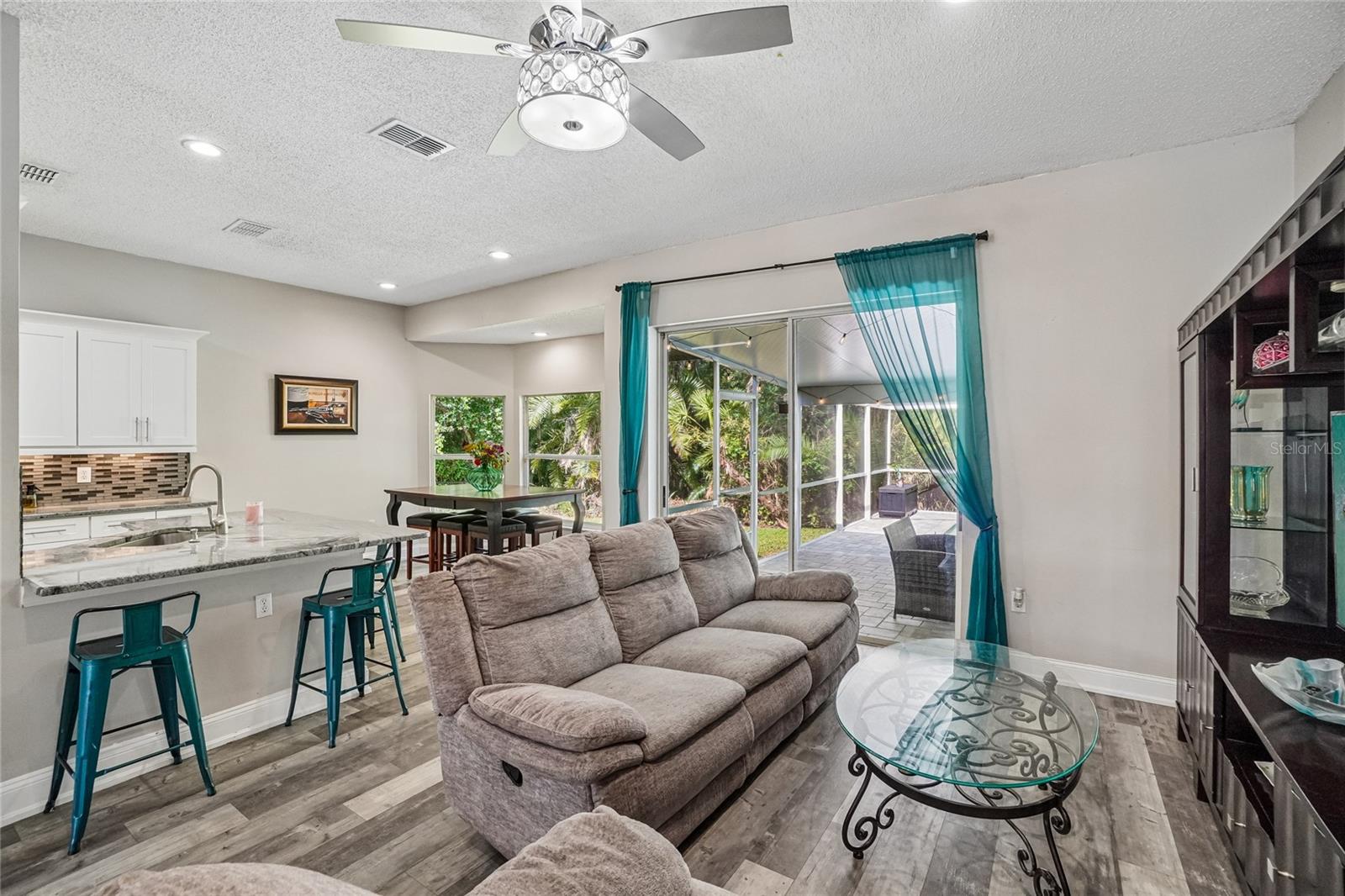 Living room / great room — Open great room with luxury vinyl plank flooring, ceiling fan, and sliding glass door to the screened lanai. Kitchen peninsula with granite countertop and tile backsplash reinforcing the open-concept layout.