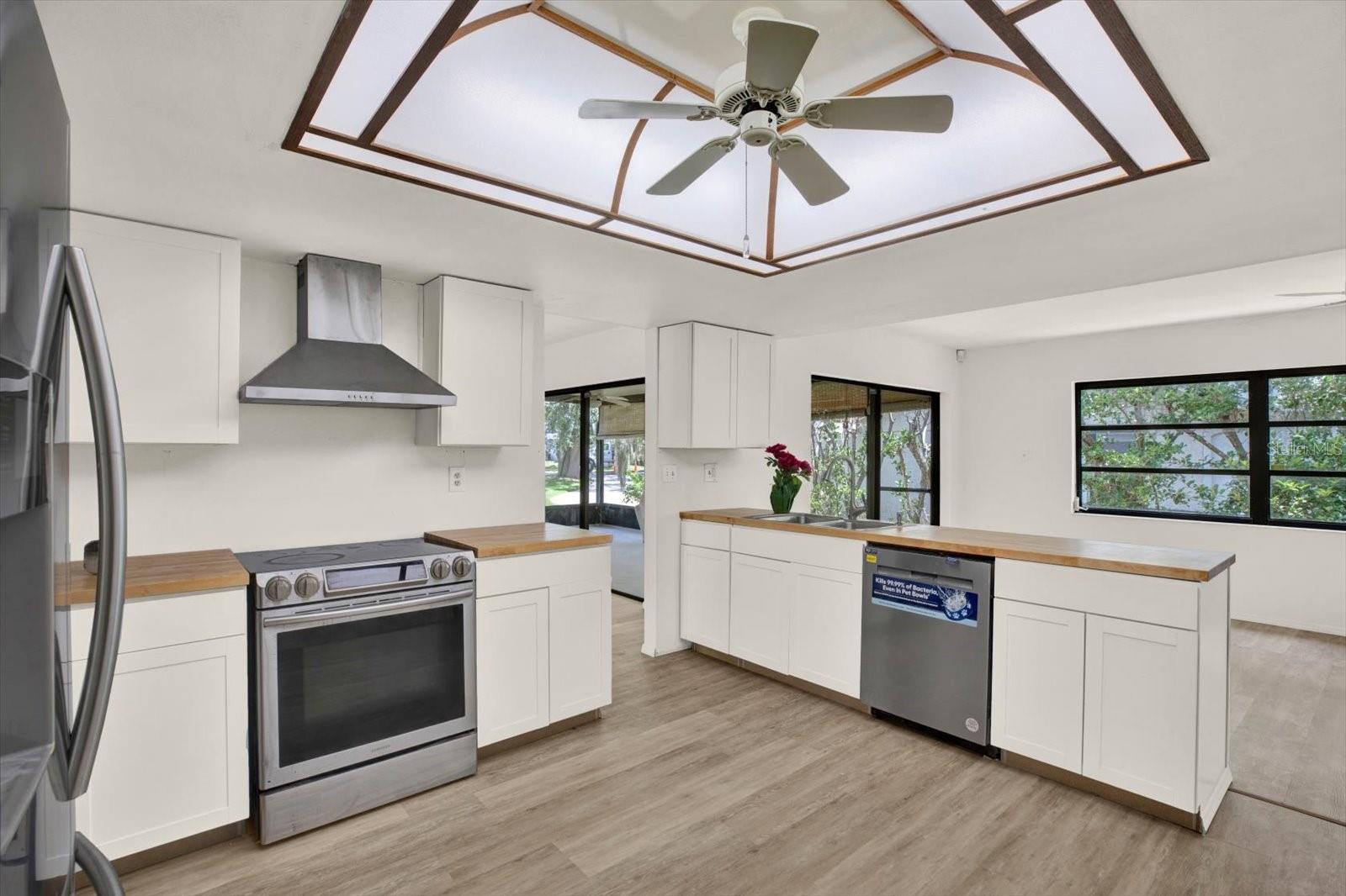 A modern kitchen featuring sleek white shaker cabinets, stainless steel appliances, a unique tray ceiling with a fan, and a view into the adjacent living space.