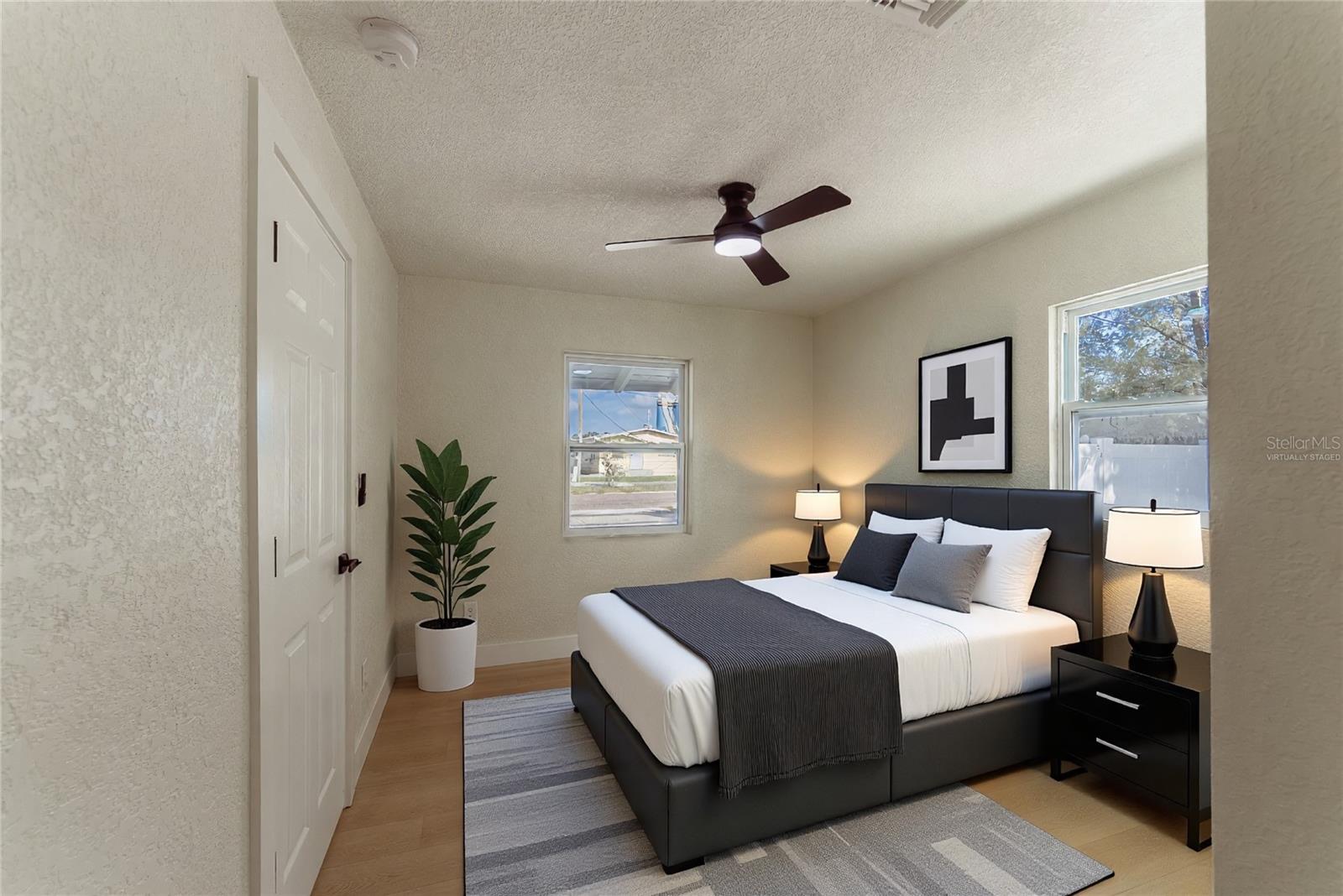 Two new impact windows flood the primary bedroom with natural light, while warm luxury vinyl flooring and a modern ceiling fan complete the space. The en suite bathroom is just steps away through the private door. (Virtually Staged.)