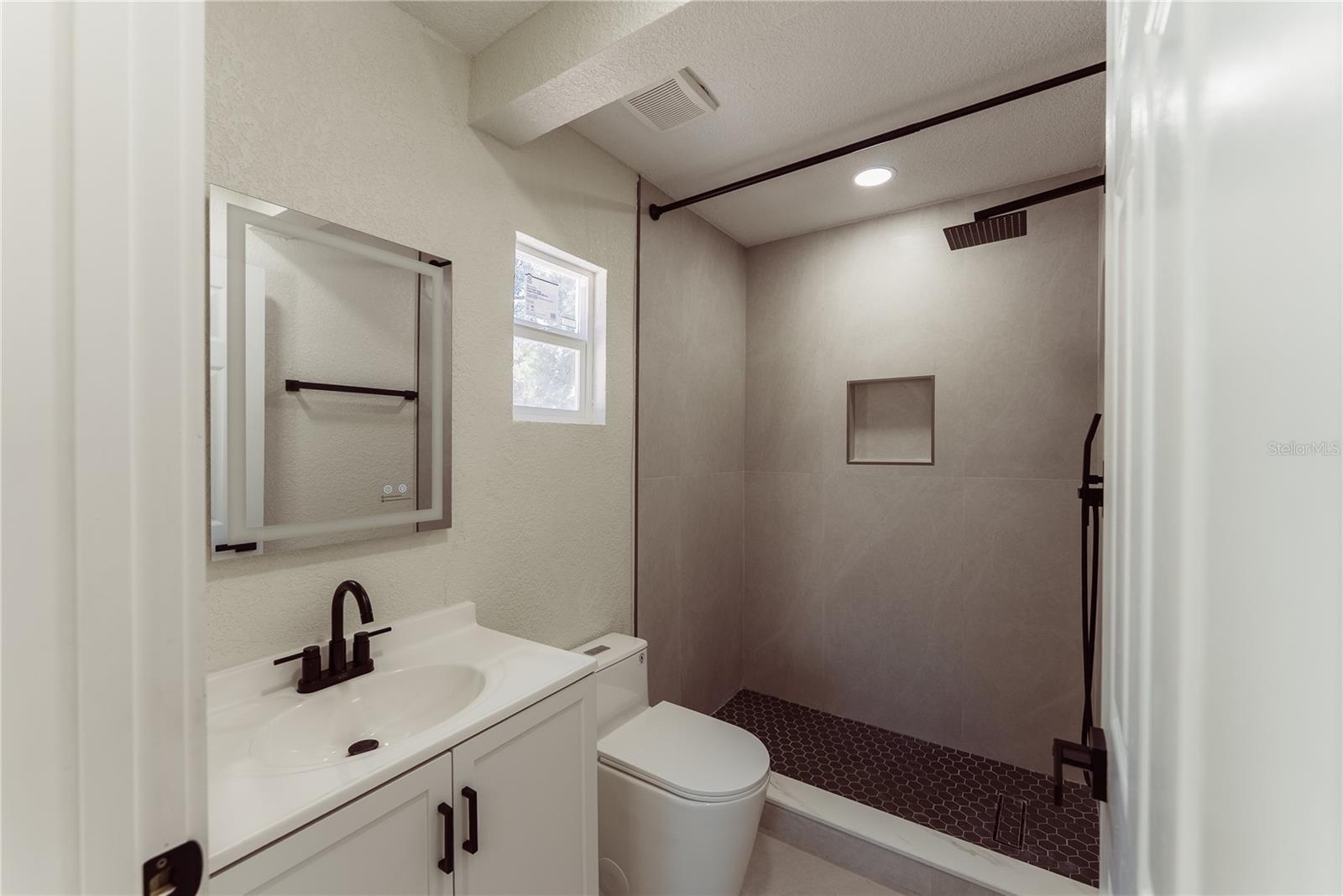 No detail was spared in this fully updated en suite, showcasing a floor-to-ceiling tiled walk-in shower with a built-in niche, rainfall showerhead, matte black hardware, and dark hex tile flooring for a sleek, modern finish.
