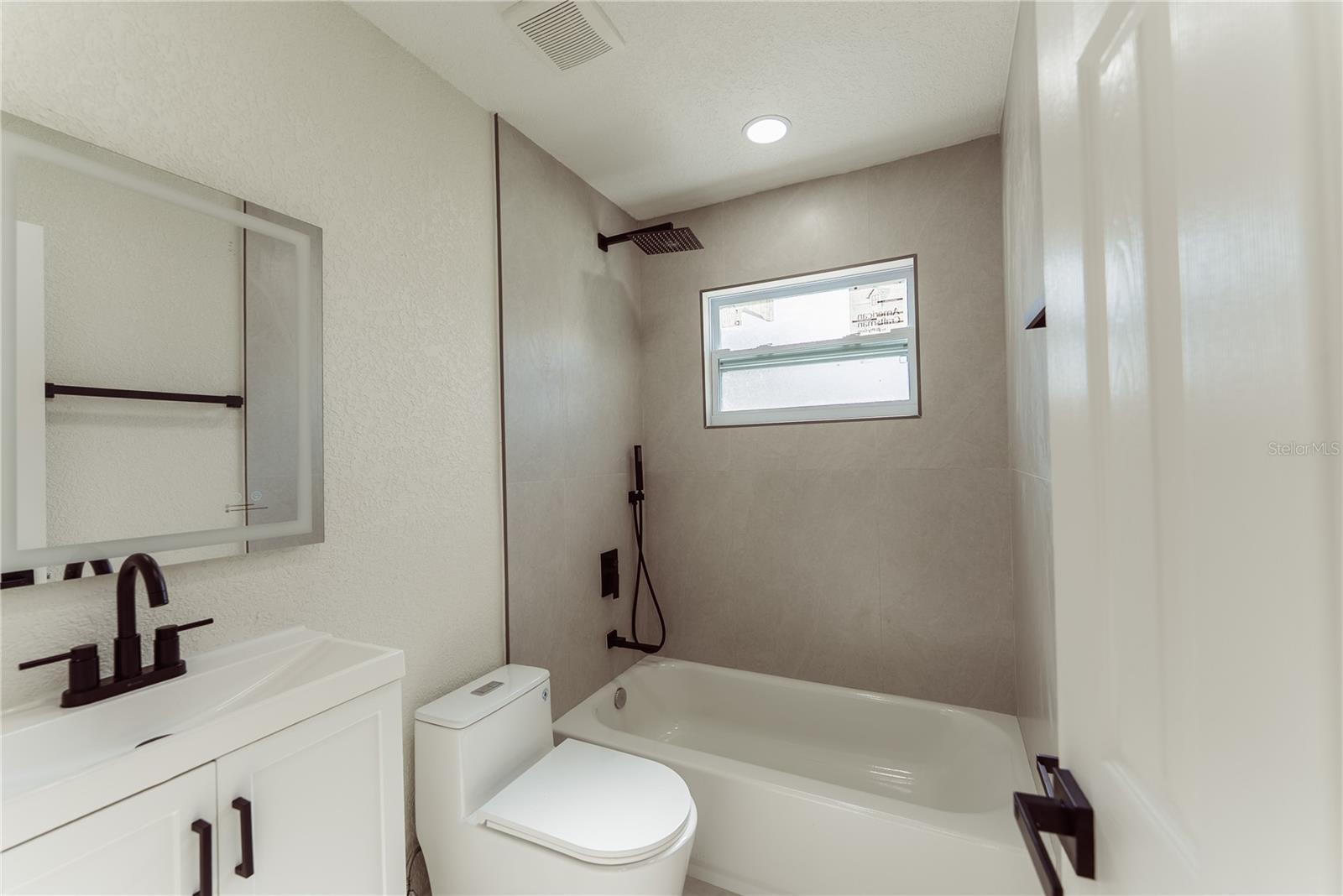 The fully updated second bathroom features a tiled tub/shower combo with a rainfall showerhead, matte black fixtures, white shaker vanity, and a natural light impact window — all brand new.