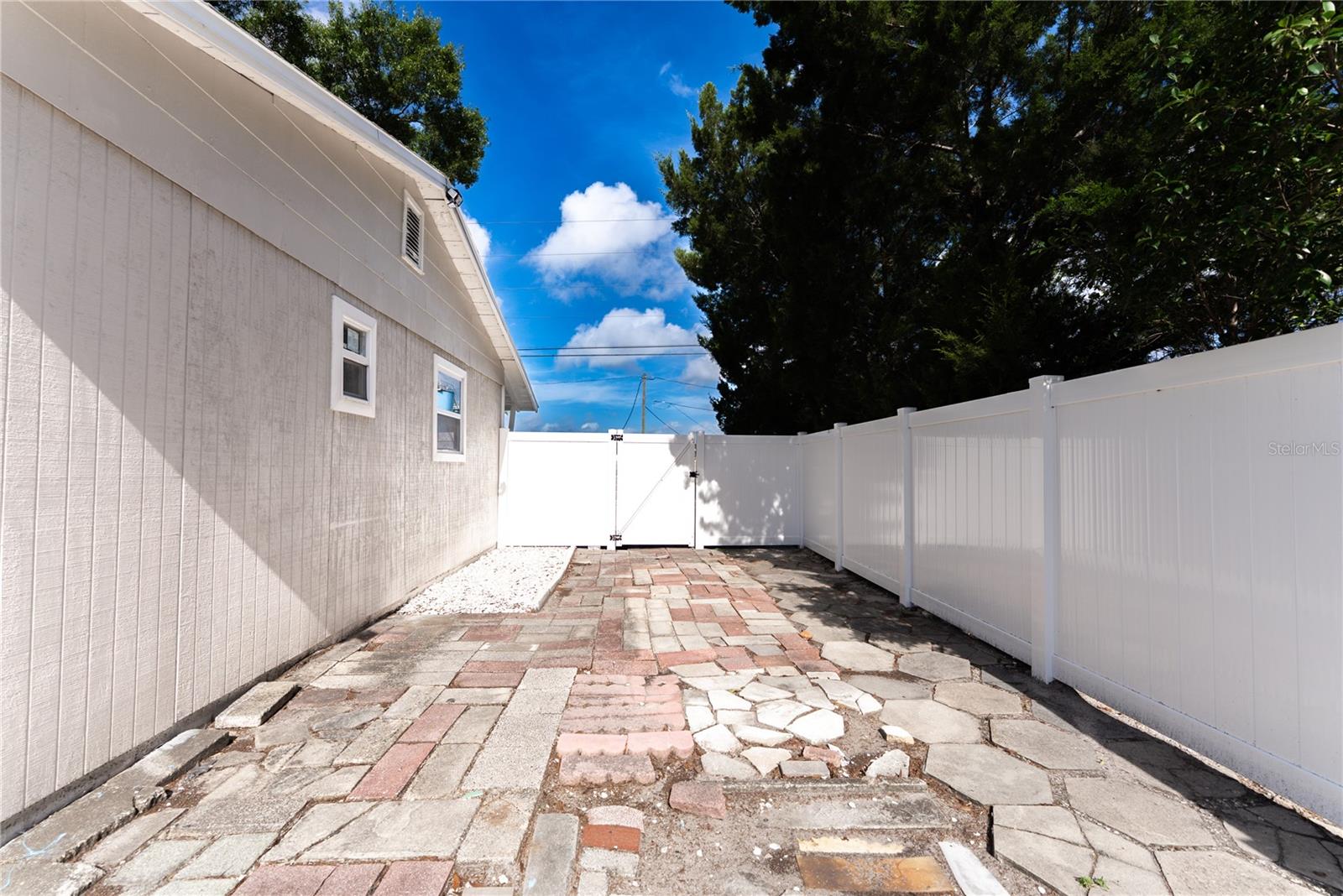 The side patio features a generous paved area ideal for use as a carport, boat storage, or additional outdoor entertaining space, with a gated white vinyl fence providing secure, private access from the exterior.