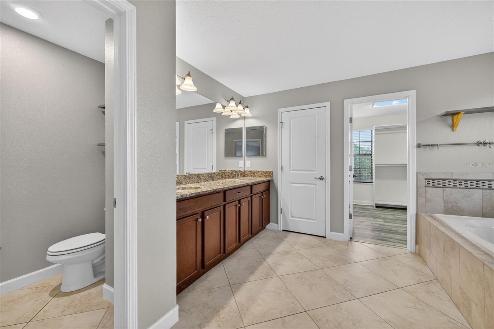 Primary En-Suite Bathroom with Dual Vanities, Granite Countertops, Garden Soaking Tub with Window, Shower Stall with Glass Frame, Private Water Closet, Linen Closet, and Entry to Custom Walk-In Closet.