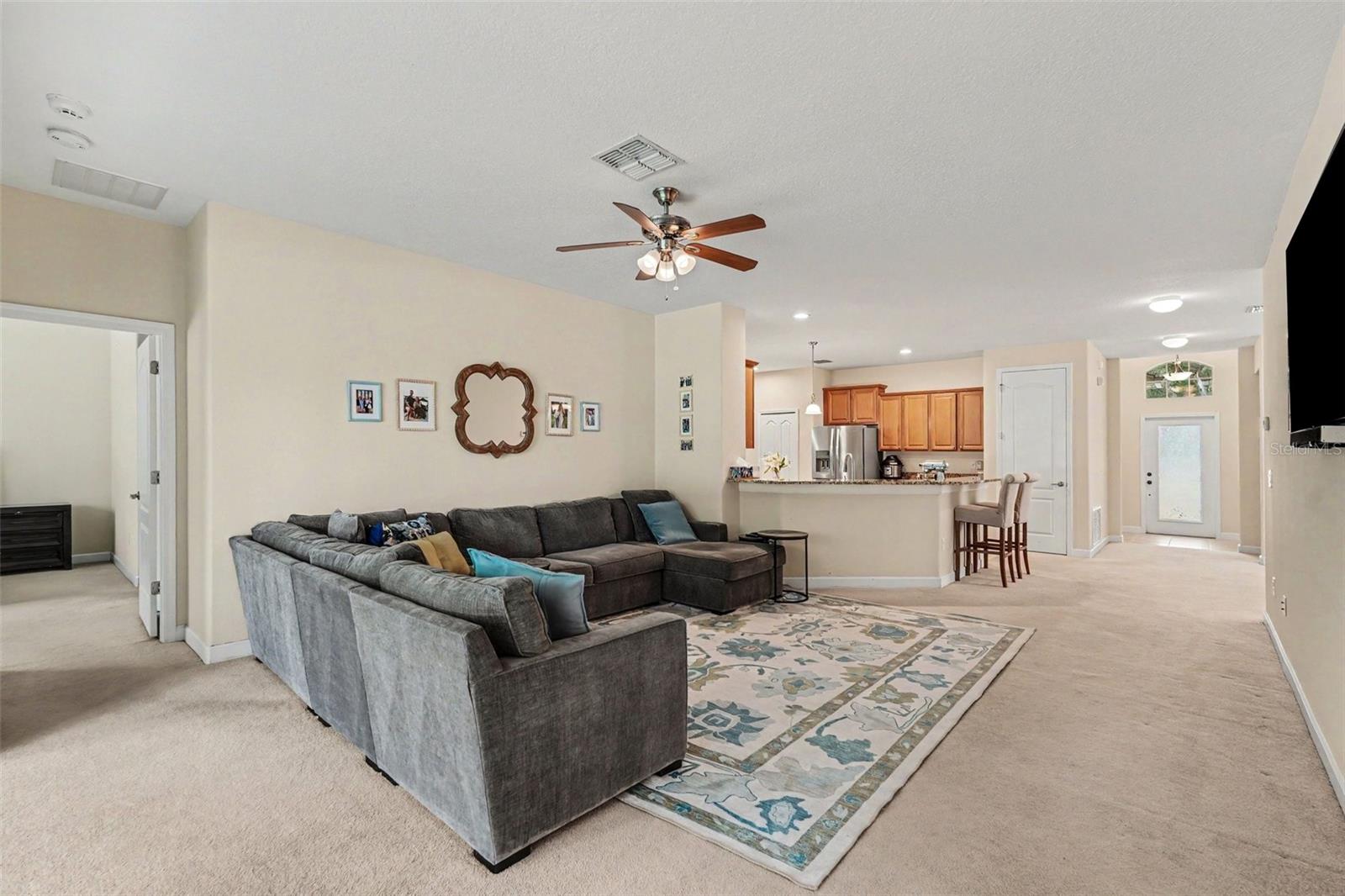 Open-concept family room with ceiling fan, and open sightline to granite kitchen peninsula, bar seating, and foyer in Tampa FL 33647. Primary bedroom suite to left.