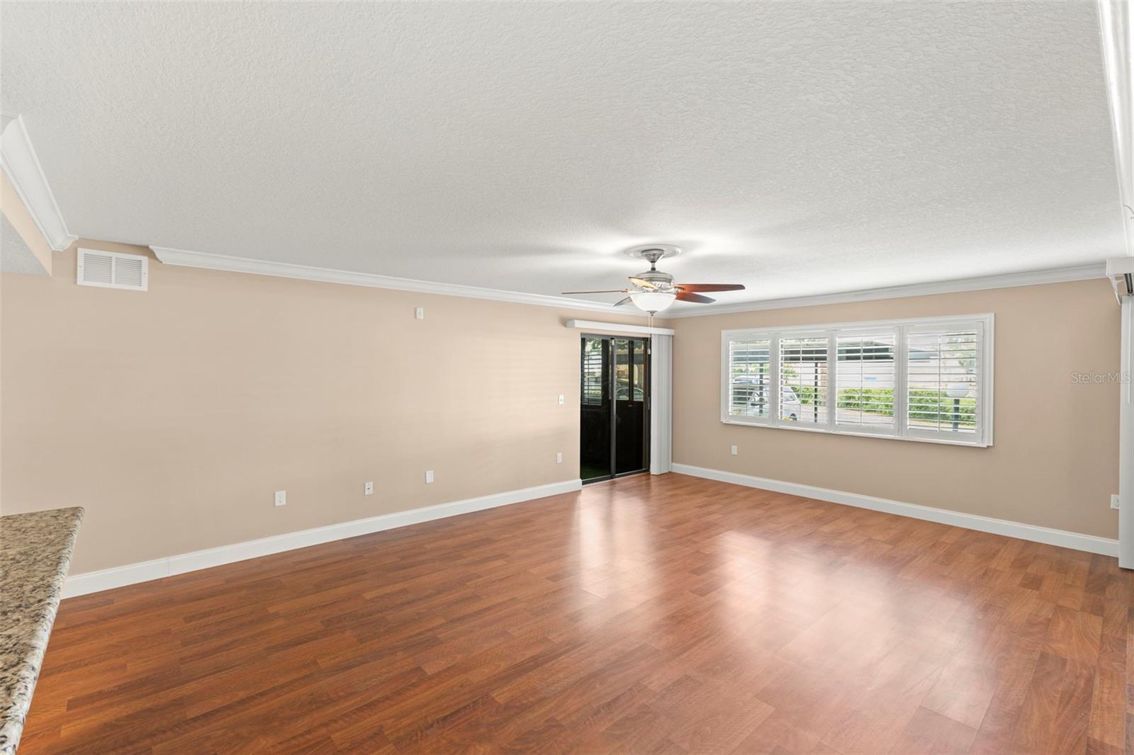 The spacious living room features wood-look flooring, crown molding, plantation shutters, a ceiling fan, and abundant natural light.