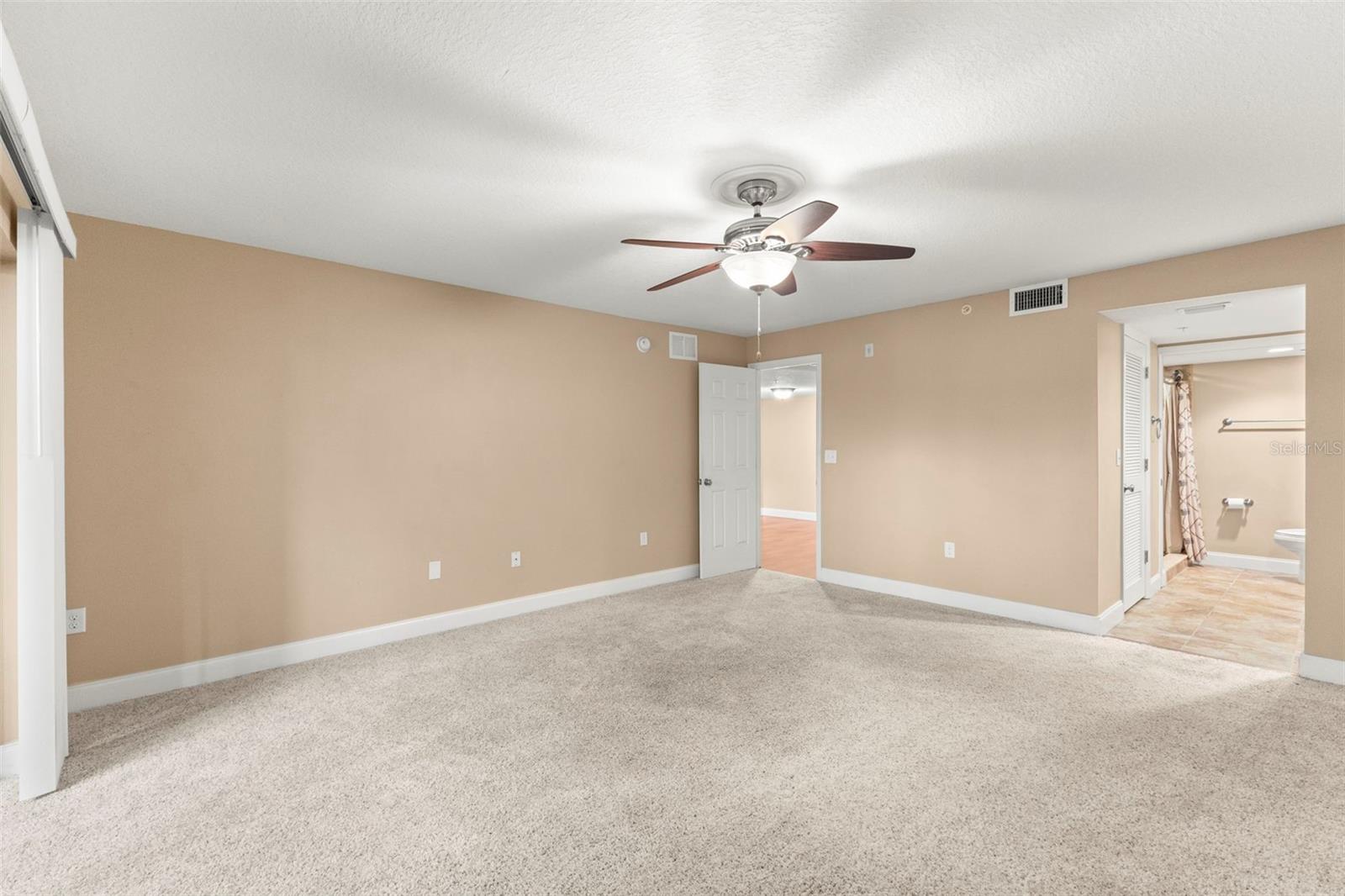 The spacious primary bedroom features plush carpeting, a ceiling fan, a neutral color palette, and direct access to the ensuite bath and walk-in closet.