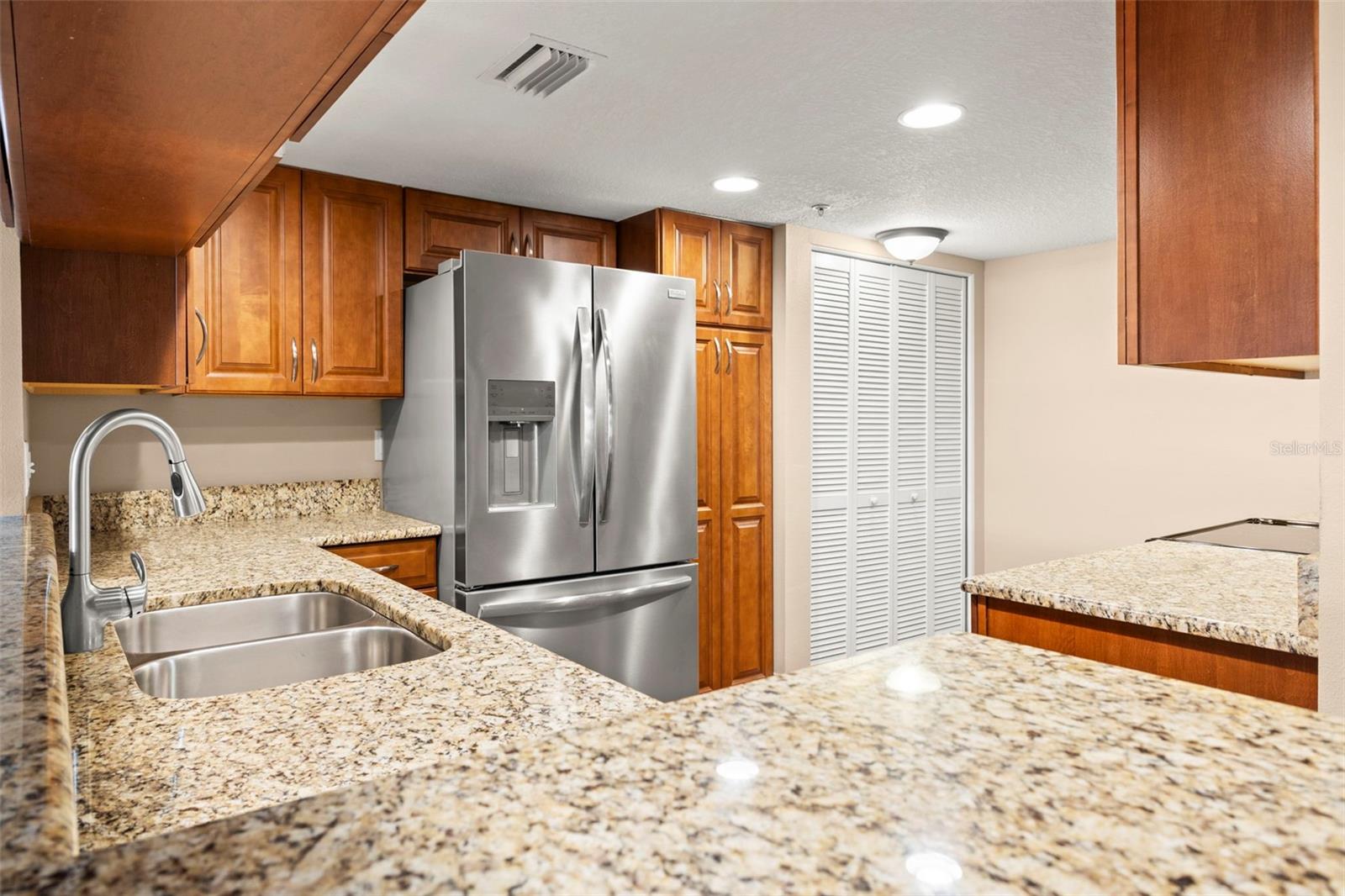 The open kitchen features granite countertops, extensive wood cabinetry, a stainless steel double-basin sink, stainless steel appliances, including a French-door refrigerator with water and ice dispensers, and recessed lighting