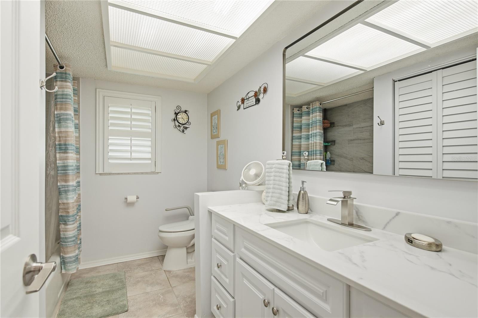 Updated secondary bathroom featuring a bright vanity with stone countertops, a large mirror, a tiled shower/tub combo, and a stylish plantation shudder window.