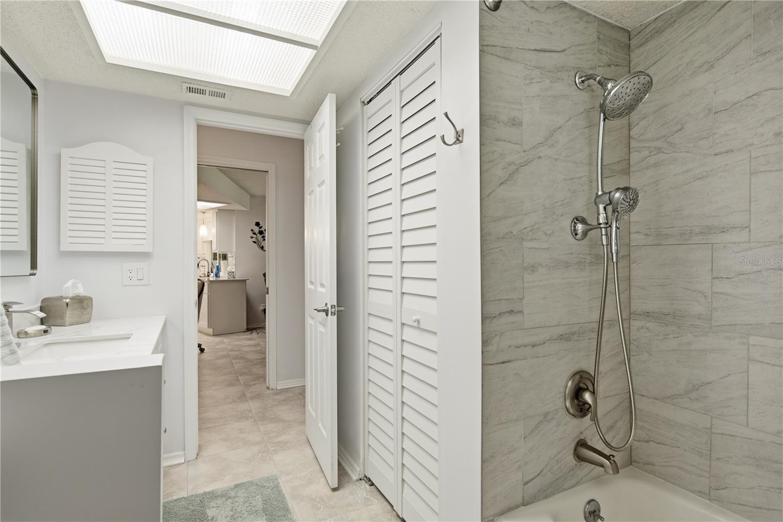 Secondary bathroom Split View highlighting an updated tub/shower combo with modern grey tile, dual shower heads, and a hallway view showing the bathroom storage closet.