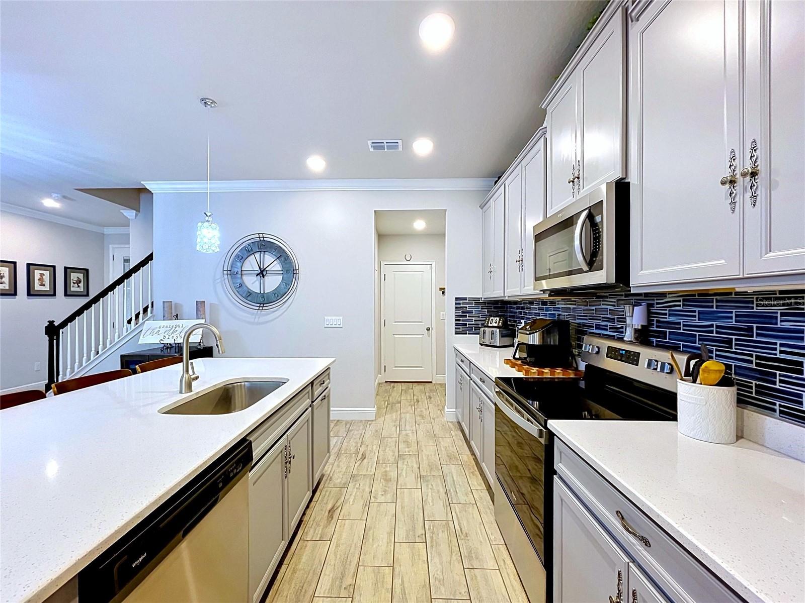 The heart of the home is this beautifully appointed kitchen featuring quartz countertops, stainless steel appliances, light gray raised-panel cabinetry, and a striking navy glass tile backsplash that adds the perfect pop of sophistication.