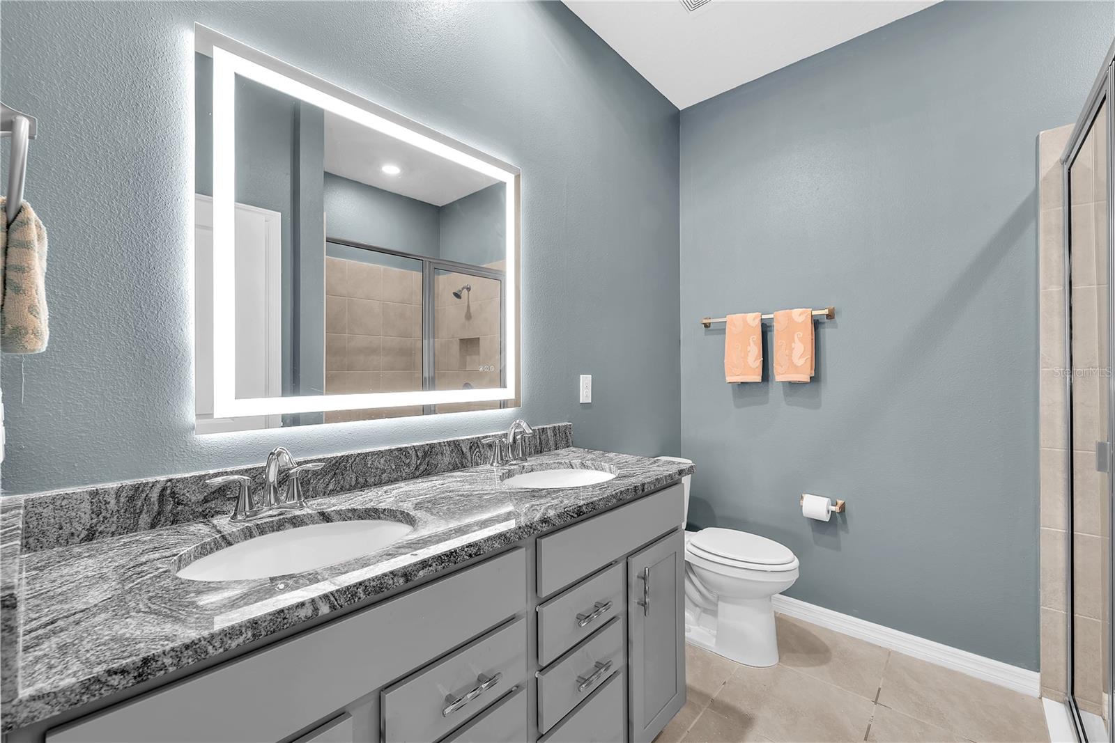 This luxurious en-suite bathroom features dual sinks, spacious vanity with granite and a touch-activated and LED lighted mirror, a large shower with built-in bench and linen closet for additional storage.