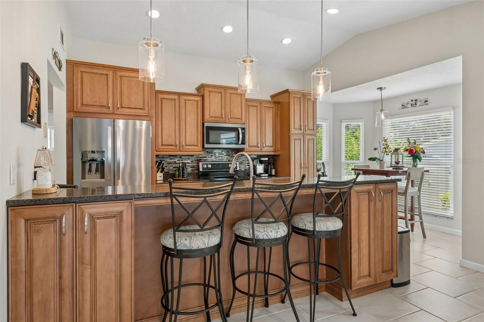 The eat-in kitchen is beautifully appointed with stainless steel appliances, granite countertops, breakfast bar seating, cabinet pullouts, soft-close drawers, and a cheerful breakfast nook surrounded by windows.