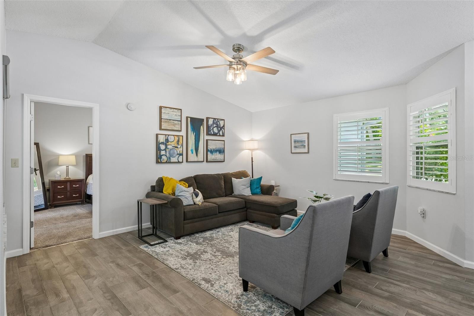 Living room with vaulted ceilings, wood-look tile floors, plantation shutters, and views out to the patio.