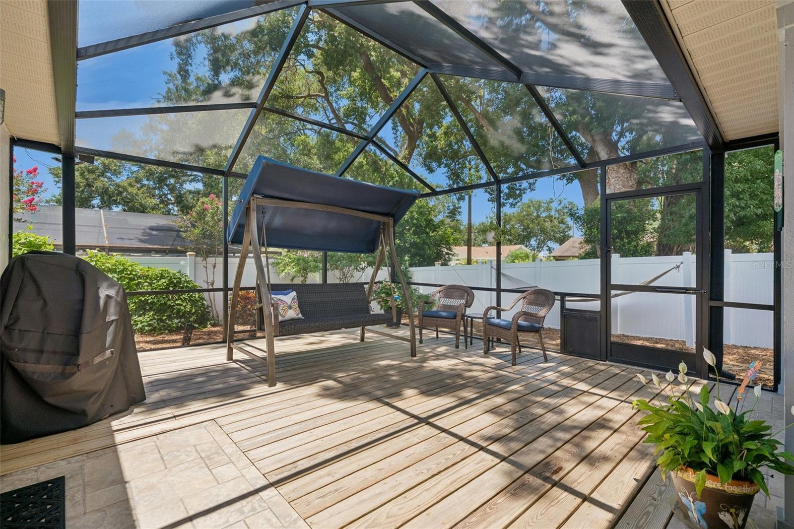 Oversized screened lanai featuring a covered seating area and wood deck, perfect for relaxing, dining, grilling, or enjoying peaceful evenings at home. Beyond the lanai, the xeriscaped backyard offers low-maintenance outdoor space with mature landscaping, tropical greenery, and a private fenced setting.